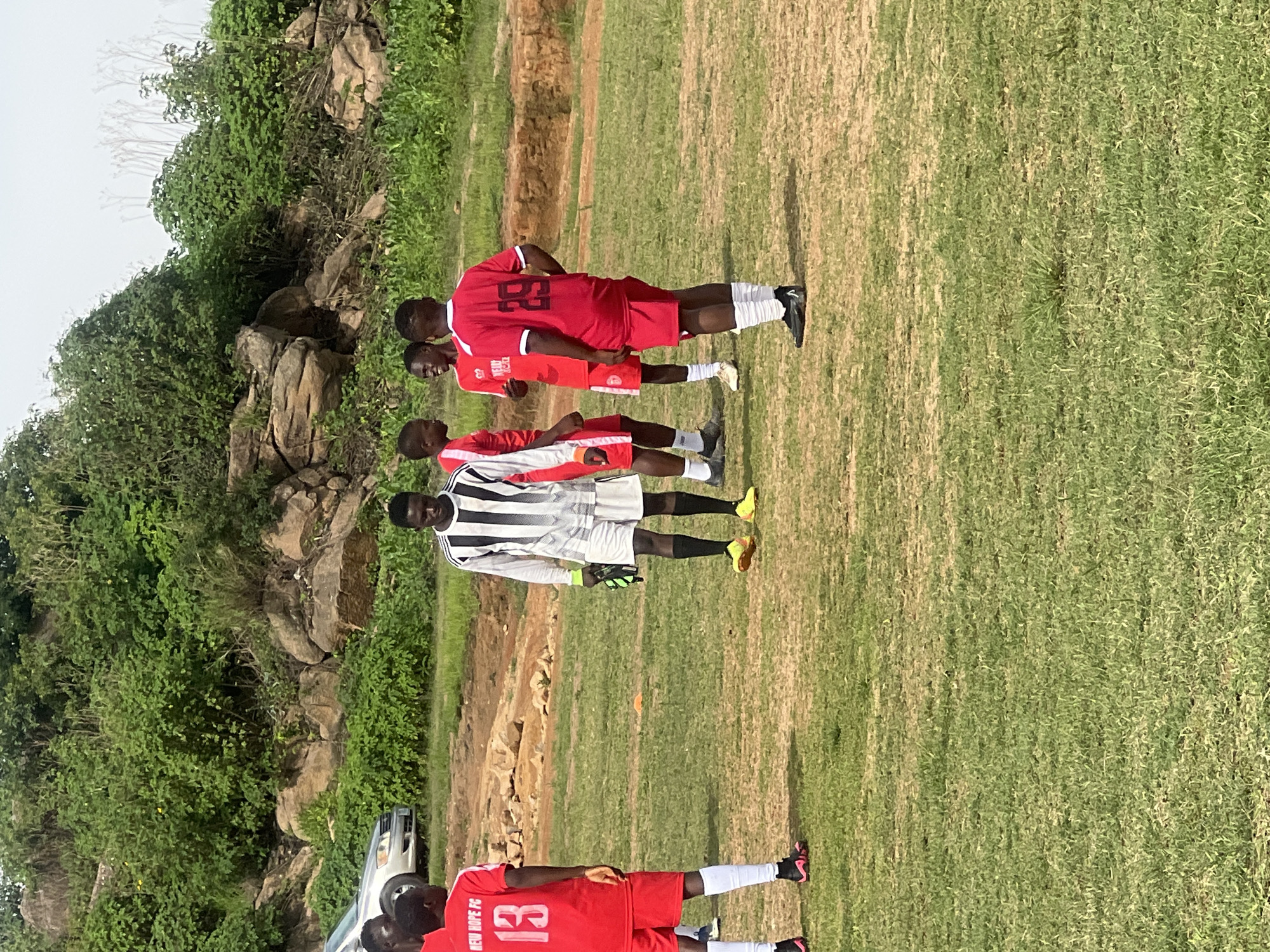 This image depicts a group of young men in athletic attire on a grassy field, possibly a soccer pitch, in Jos, Nigeria. The individuals are dressed in red and black and white striped jerseys, suggesting a team or practice setting. One person wears a red jersey with the number 13, and another sports a red jersey with the number 29. The background features rocky terrain and lush green vegetation, indicative of an outdoor environment in Jos, a city known for its hilly topography and vegetation. A silver car is partially visible on the left side of the frame. The ground is uneven, with patches of dry earth and green grass. The overall scene suggests a casual sports activity or training session in a natural setting.