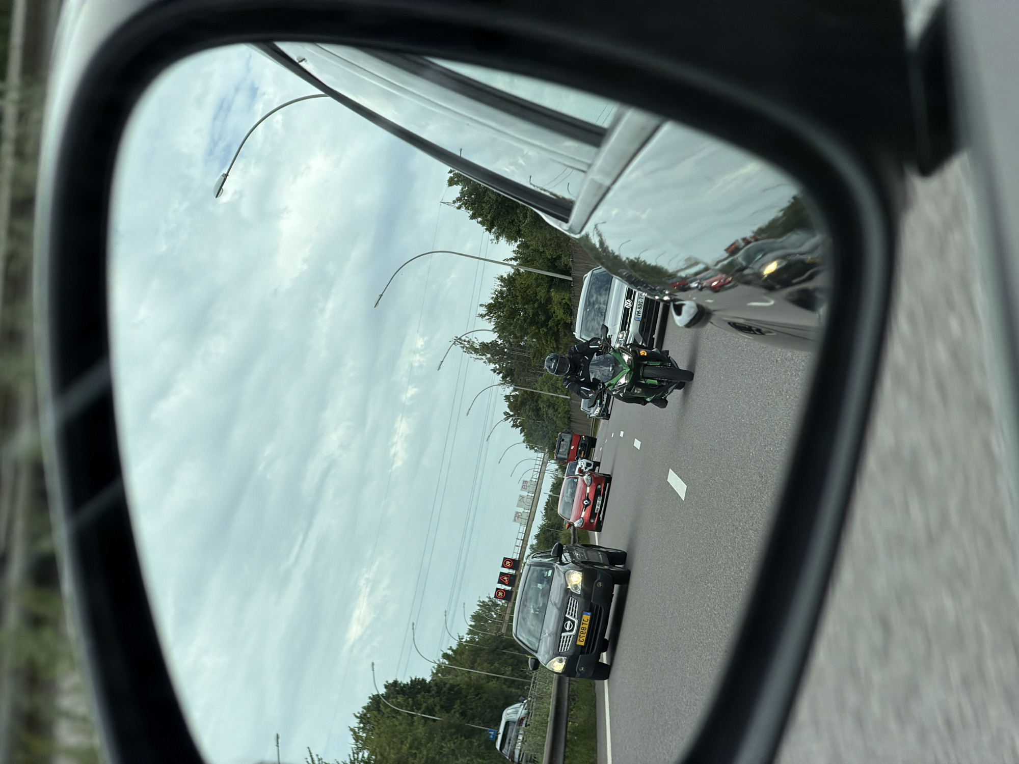 This is a photograph taken from the perspective of a driver's side mirror, showing traffic on a highway in Mondercange, Luxembourg. The mirror reflects a cloudy sky with streaks of sunlight breaking through. On the highway itself, a green Kawasaki motorcycle with a rider in a black helmet and racing suit is in motion. Behind the motorcycle, a silver Volkswagen hatchback is visible. Further ahead, a black Nissan Qashqai is in the left lane, and a red Renault Clio is in the right lane. A red truck is also visible in the distance. The highway is flanked by trees and lampposts. The image is dated June 27, 2023, at 11:44 AM.