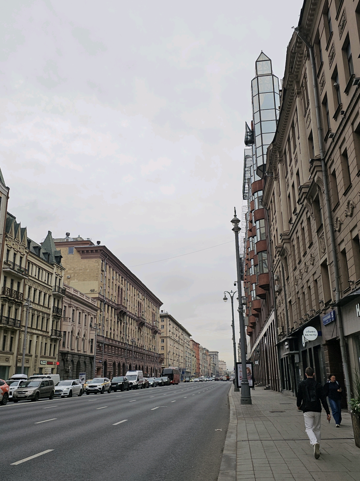 This is a medium-shot, eye-level, outdoor, daytime photograph taken from the sidewalk looking down a street in Moscow, Russia. The sky is overcast with pale grey clouds. The street is a wide, multi-lane asphalt road with white lane markings. The road is lined with cars parked on the right side and driving on the left. Buildings line the right side of the street, featuring neoclassical architecture with ornate facades, arched windows, and balconies. A modern glass tower with a pointed roof is visible above some of the older buildings on the right. On the left side of the street, more buildings with similar architectural styles are visible, receding into the distance. Pedestrians are walking on the sidewalk. One person in a black jacket and white pants is walking away from the camera, while another person in a dark jacket and jeans is walking towards the camera. A street lamp stands on the sidewalk. The overall impression is an urban street scene in Moscow on a cloudy day.