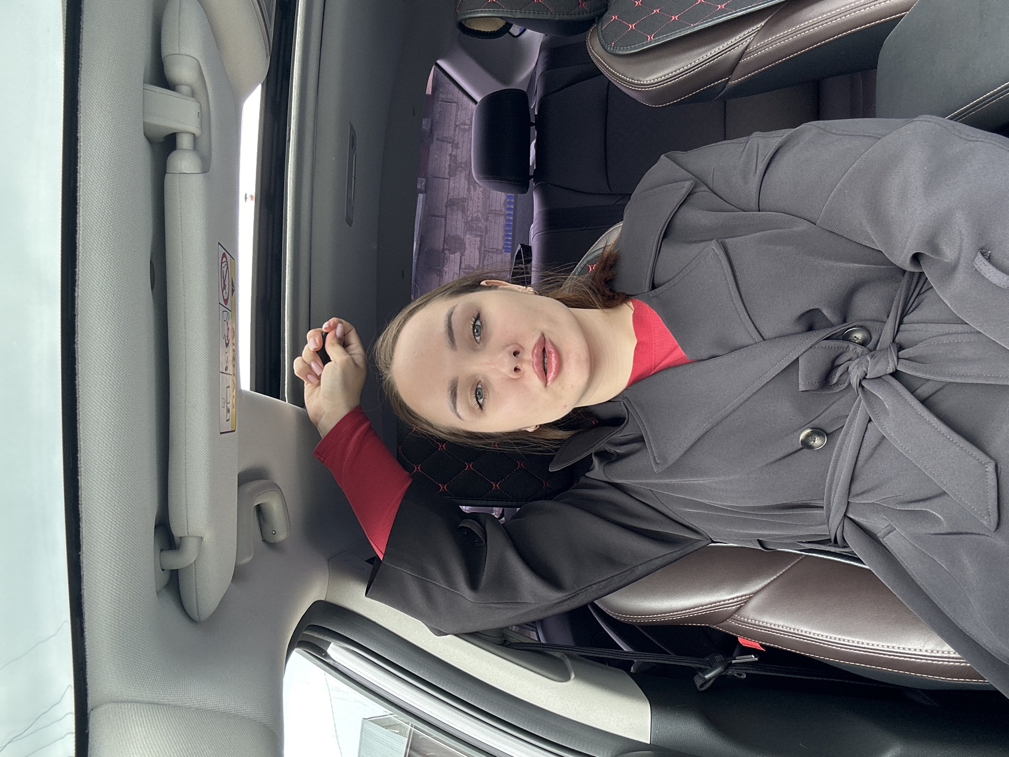 A woman lies sideways in the backseat of a car. She wears a red shirt and a dark gray trench coat tied at the waist. Her head rests near the window, and she looks at the camera. The interior of the car includes dark leather seats with red patterned headrests, light gray ceiling and door panels, and a sun visor. The location is in Chaikovsky, Russia, based on the car's origin, a common car model in the area, and the woman's appearance, which aligns with local demographics.