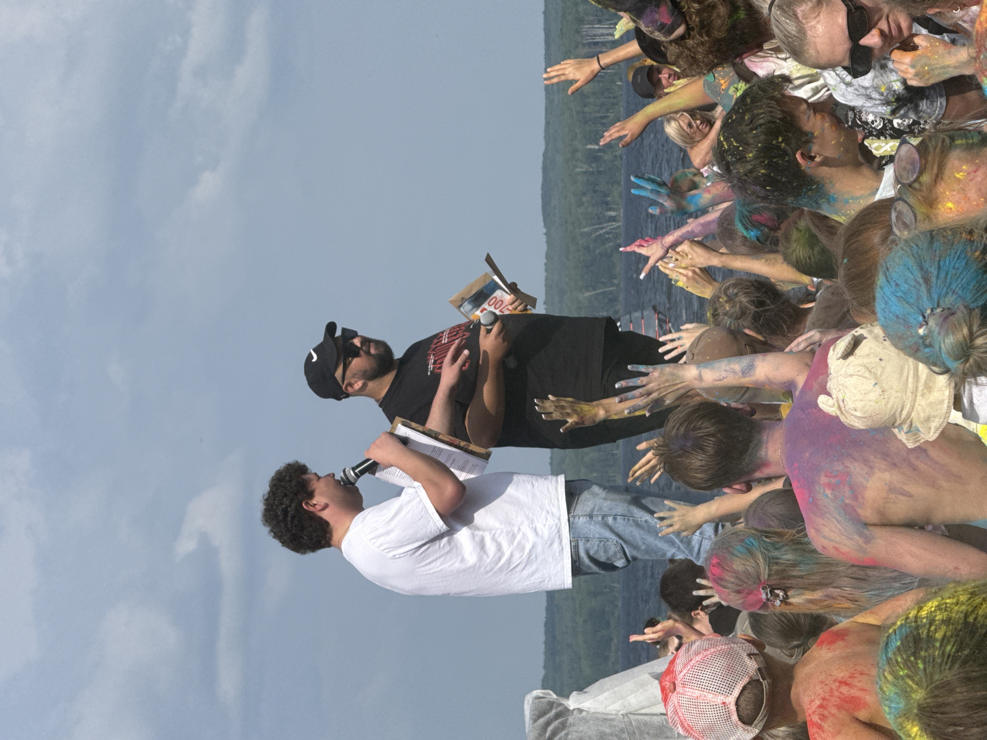 The image shows an outdoor gathering, likely a festival, with a crowd of people, many of whom are covered in colorful powder, suggesting a Holi celebration. Two men stand on a slightly elevated platform in the background, addressing the crowd. The man on the left, wearing a white t-shirt and jeans, holds a microphone and a piece of paper. The man on the right, dressed in a black t-shirt and a black baseball cap, holds a pamphlet or magazine. The background features a body of water and a treeline, indicative of a natural setting near a city. This scene is likely located in the city of Chaikovsky, Russia, given the reference information.