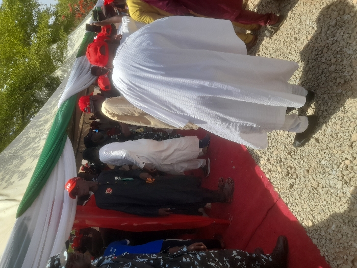 The image shows a group of people standing under a white tent with a green stripe. The tent is decorated with red fabric. The people are standing on a gravel surface, with a red carpet leading into the tent. There are trees and greenery behind the tent. The group of people are gathered outside a building located in Gusau, Nigeria (Latitude: 12.156, Longitude: 6.71). The scene is likely a public gathering, as many people are present and there are several people wearing traditional Nigerian clothing. There is one man wearing a white robe, and another man wearing a black robe. Most people are wearing red hats. The image shows a man in the white robe standing in the foreground, while many other people are blurred in the background.