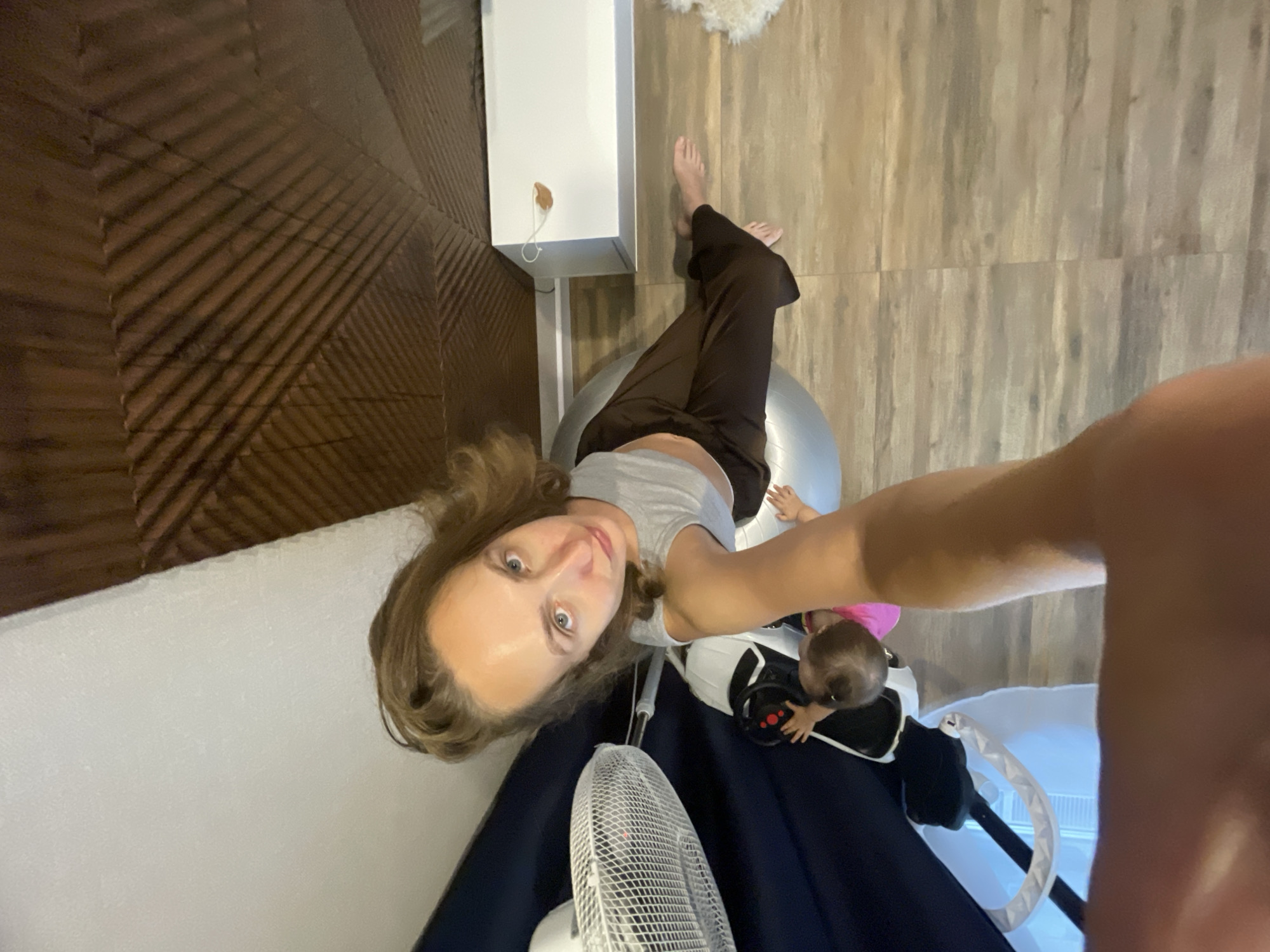 A woman is sitting on a large exercise ball, her legs are bent, and her feet are flat on the floor. She is wearing a grey crop top and brown pants. Next to her, a baby in a pink onesie is reaching out. Behind the woman, there is a white fan and a dark blue surface. The floor is made of wooden planks. The ceiling has a patterned texture and appears to be angled. In the background, a white rectangular object is visible. The overall scene suggests an indoor setting, possibly a home.