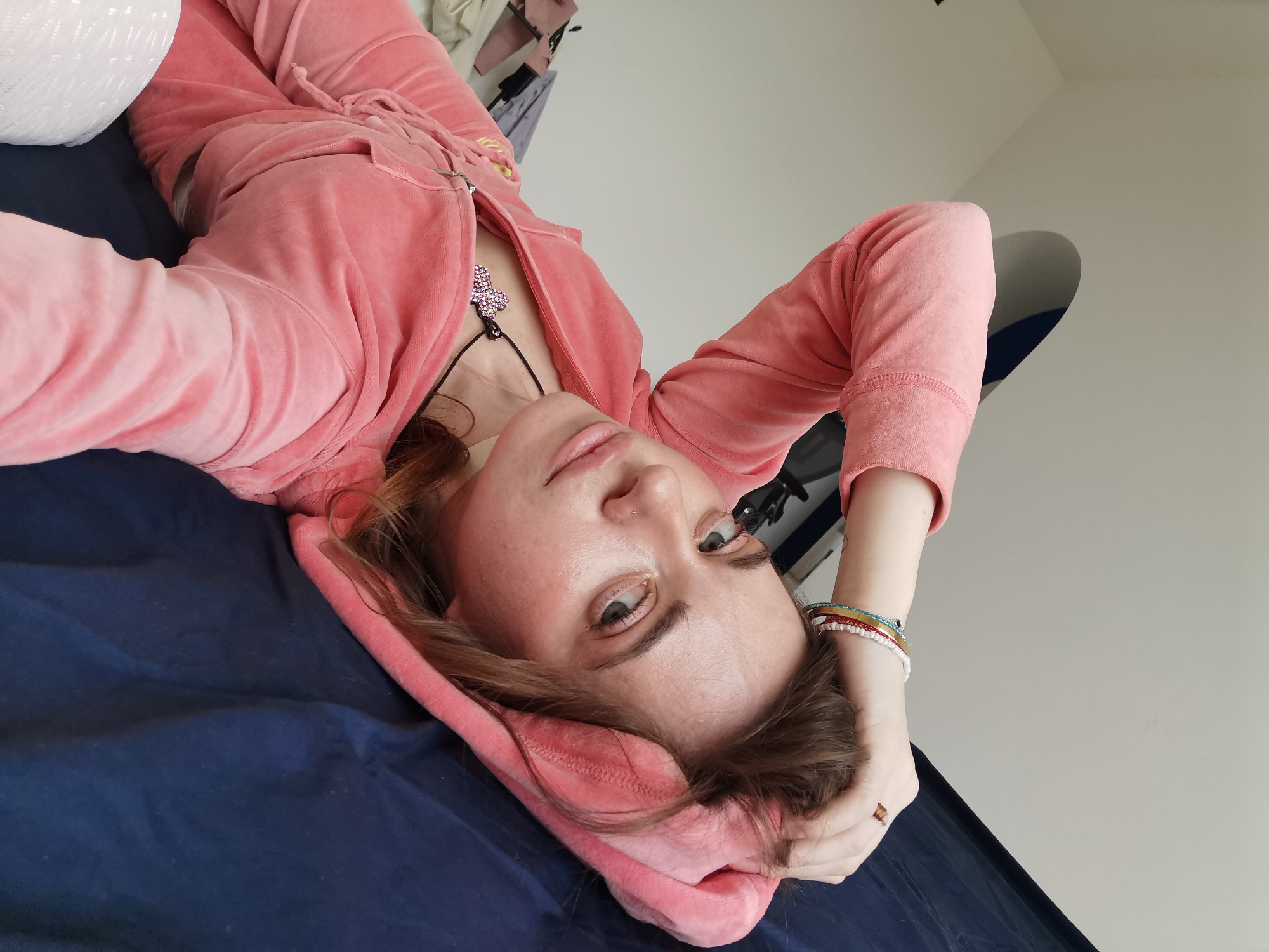 A woman wearing a pink velour tracksuit is lying on a dark blue surface, possibly a bed or couch. She is looking directly at the camera. Her head is tilted to the side, and one hand is resting behind her head. She is wearing a necklace with a heart pendant. The background shows a white wall. There is no evidence to suggest the location is Moscow, Russia.