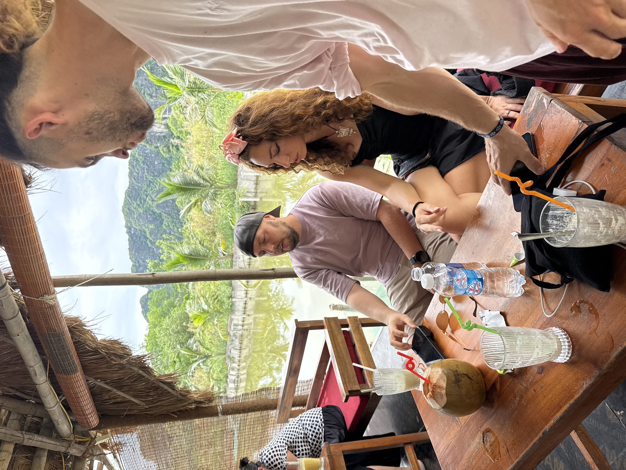 Three people are seated at a wooden table in an outdoor cafe with a thatched roof and a view of a calm body of water and lush green mountains. On the table, a whole coconut with a straw, two glasses with straws, and a bottle of water are visible. A man in a pink shirt and a baseball cap is looking down at his phone. Next to him, a woman with curly brown hair, wearing a black dress and a pink flower accessory in her hair, is looking downwards. A third person, wearing a pink shirt, is partially visible on the right, facing away from the camera. In the background, palm trees and other foliage line the water's edge. The scene suggests a relaxed outdoor dining or drinking experience.