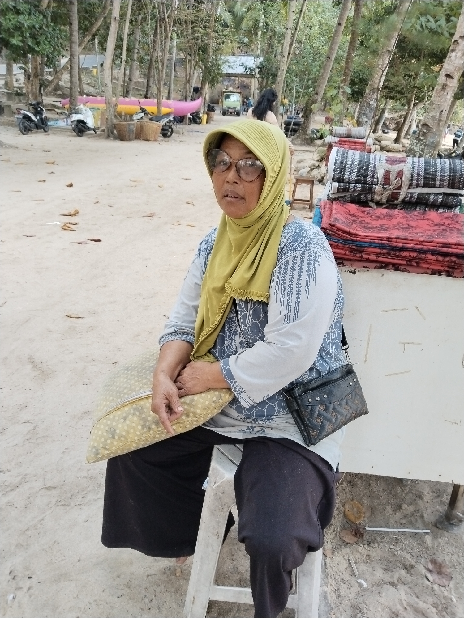 A woman wearing a yellow hijab and glasses sits on a white stool on a sandy surface near a line of trees. She is holding a light-colored pillow with a dotted pattern. She wears a light gray long-sleeved shirt with a blue and white pattern and black pants. A small black purse with a diamond pattern and silver studs is slung across her body. Behind her, stacked mats of various colors are visible. In the background, several motorcycles and a banana boat are parked on the sandy ground. A few people are visible in the distance among the trees, and a green truck is parked further back. The scene appears to be a beach or coastal area in Prigi, Indonesia.