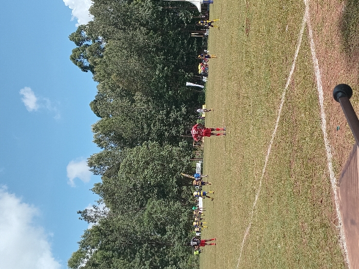 The image depicts an outdoor sports field situated in Meru, Kenya, under a blue sky with scattered white clouds. A dense line of tall green trees forms the background, bordering the playing area. The field itself is covered in green grass, marked with distinct white lines indicating boundaries or play zones. Approximately fifteen individuals, presumed to be players, are distributed across the field, wearing various sports uniforms. One prominent figure, located centrally, wears a red jersey with the number "12" visible on the back and red shorts, standing with hands on hips, facing away from the camera. Another player in a black jersey with the number "9" and red shorts is positioned in the mid-left. Other players are dressed in yellow and black, green and yellow, or blue jerseys. In the far background, adjacent to the tree line on the right, two canopy structures, one white and one blue, are discernible. The individuals appear to be engaged in a sporting activity, with most figures in static or low-activity poses. A dark, cylindrical object and a brown, textured surface are partially visible in the immediate bottom right foreground.