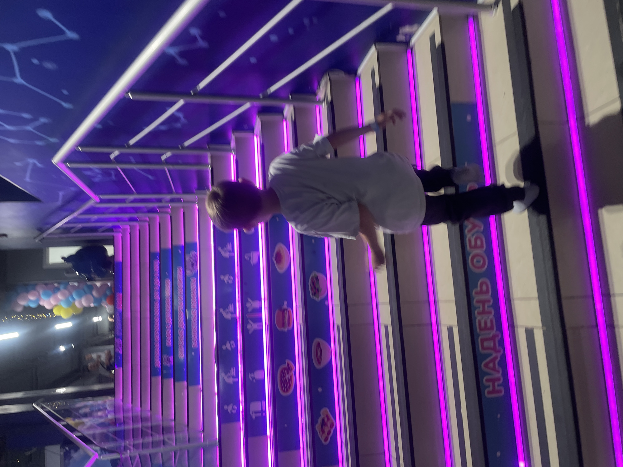 Here's a detailed description of the image:
**General Scene:** The image appears to be taken indoors, likely at a public venue in the city of Sochi, Russia. The environment features stairs with vibrant purple neon lighting, accompanied by informational boards and a decorative ceiling. The stairs have the Russian words "НАДЕНЬ ОБУВ" written on them.
**People:** There is one individual in the frame, a person with light-colored hair. They are wearing a light-colored shirt, dark pants, and appear to be walking on the stairs.
**Objects:**
* Stairs with purple neon lighting on the side of each step
* Informational boards with images
* Decorated ceiling with constellation-like patterns
* Balloons in the background, suggesting a festive or celebratory atmosphere.
**Actions/Interactions:** The individual is walking on the stairs. The informational boards seem to be promoting food items, given the presence of food-related images.
**Environment:** The setting is indoors, with modern and vibrant design elements. The lighting is predominantly purple, creating a lively atmosphere. The presence of stairs and informational boards implies a public or commercial space, possibly a market or exhibition in Sochi.