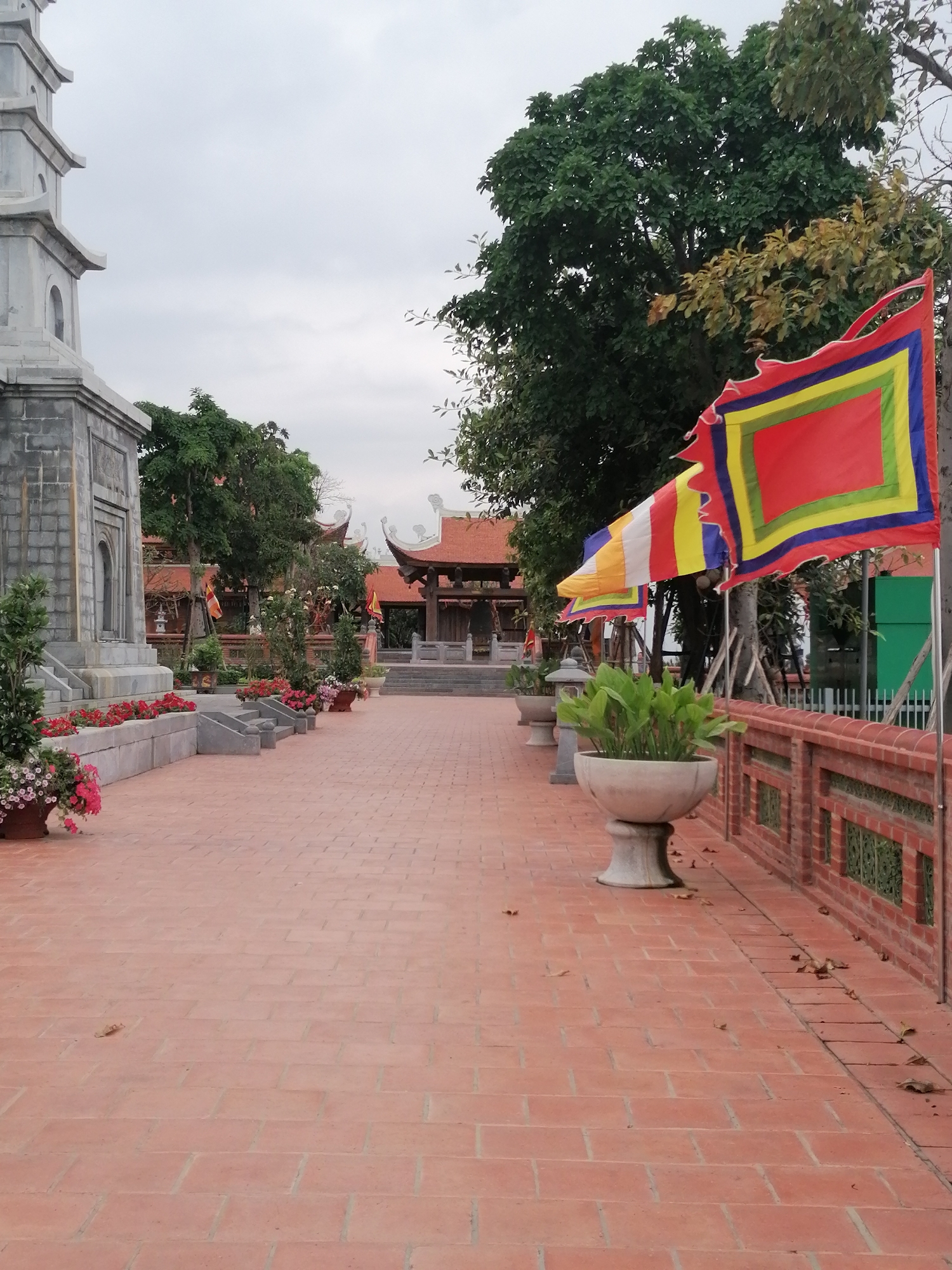 The image depicts a brick-paved walkway in Hong Gai Ward, Vietnam. To the left stands a gray stone pagoda-like structure with plants and flowers in pots along the base. Red-roofed buildings are visible in the background. Along the right side of the walkway is a red brick wall with decorative openings. Potted plants line the top of the wall. Several colorful Buddhist flags are mounted on poles and stand near the wall. Fallen leaves are scattered on the brick pavement.