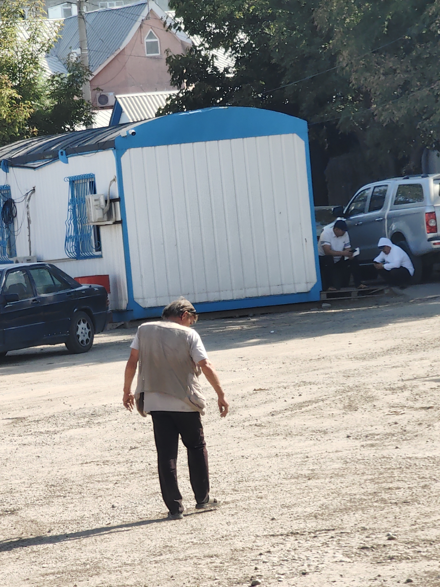 In Shymkent, Kazakhstan, on a sunlit day, a man wearing a beige vest over a white t-shirt and dark trousers walks away from the camera across a gravelly, dusty lot. To his left, a dark-colored sedan is parked. In the background, a portable white structure with a bright blue trim stands prominently. Two individuals are seated near the entrance of this structure; one wearing a blue cap and white shirt, the other in a white hooded garment. A silver pickup truck is parked behind them. The environment suggests an outdoor industrial or construction site, with trees and buildings visible further in the distance.