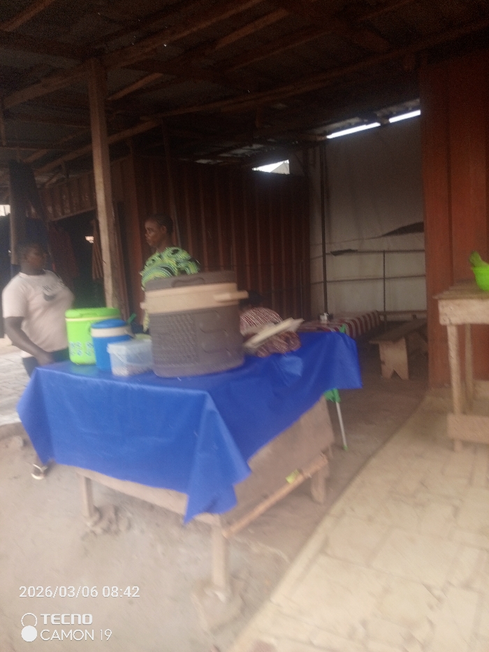 The scene depicts a semi-enclosed commercial or service area in Nsugbe, Nigeria. The structure features corrugated metal walls, some appearing brownish-red, and an exposed wooden beam roof framework. In the foreground, a wooden table is covered with a bright blue tablecloth. Positioned on this table are a lime green cooler, a blue and white lidded cylindrical container, a clear rectangular plastic container, and a large, dark-colored insulated cooler with a lid.
Two adult females are visible. On the left, a woman wearing a light-colored short-sleeved top with a dark emblem stands beside the table. In the center-right background, behind the table, another woman wearing a green and black patterned top is present. A third individual, partially obscured by the blue tablecloth and the large cooler, can be seen sitting or stooping behind the table, wearing brown and white striped clothing.
In the background, a light-colored wooden bench is situated against a wall. To the far right, a simple wooden stand holds a small, lime-green plastic bucket. The ground surface is light-colored concrete or pavement. Natural light illuminates the area. The image carries a timestamp of 2026/03/06 08:42.