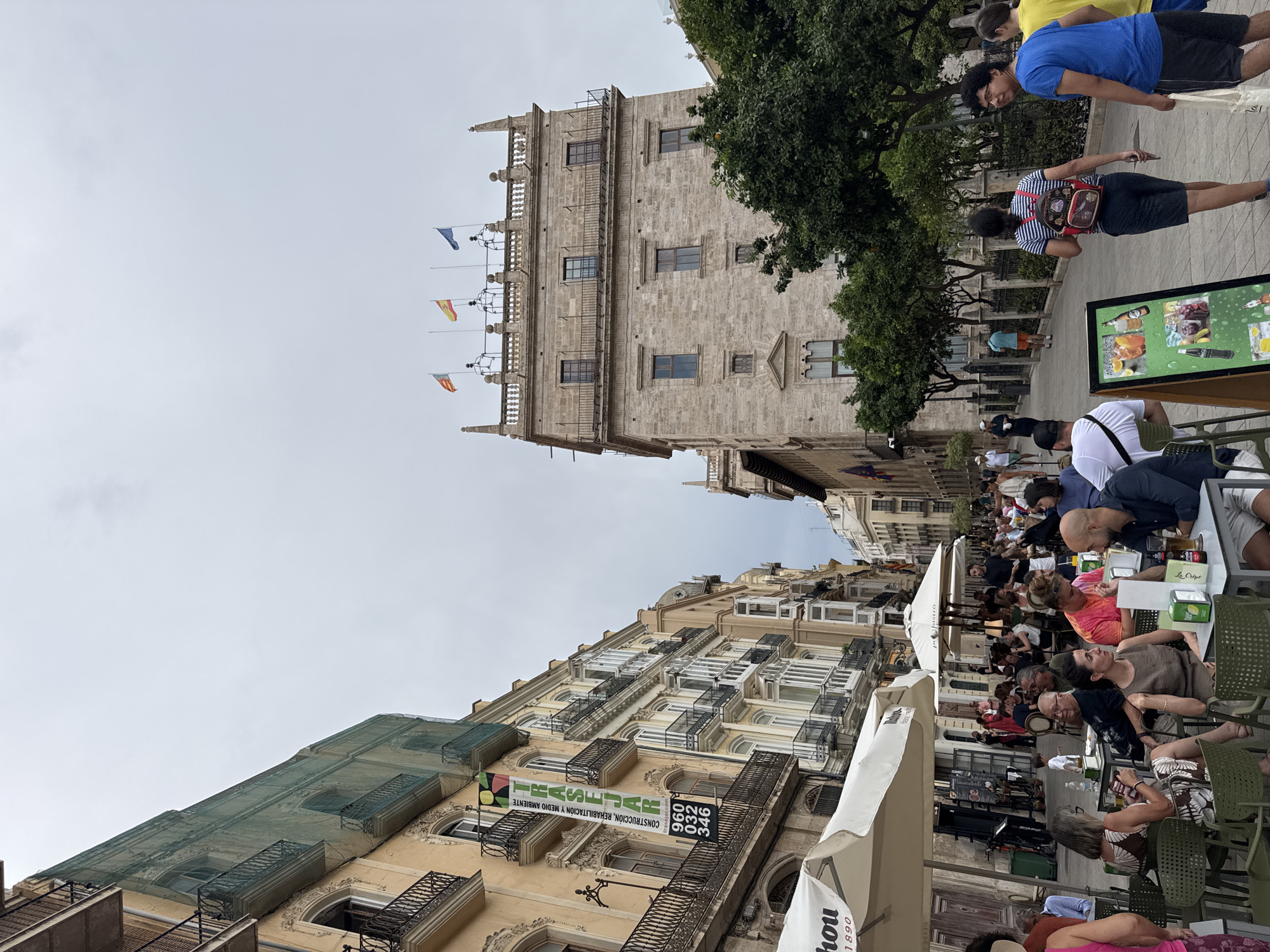 The image depicts a street scene in Valencia, Spain, likely in the vicinity of the Silk Exchange (La Lonja de la Seda). On the left, buildings with balconies and awnings line a narrow street. A banner advertising construction services hangs from one building. In the foreground, several outdoor cafe tables are occupied by people.
The focus shifts to a grand stone building in the background, featuring multiple stories, windows, and a balustraded roofline adorned with flags. This building resembles the architecture of La Lonja. Trees are planted in front of this structure.
People are engaged in various activities: walking, sitting at cafes, and conversing. The atmosphere appears casual and social. The sky is overcast, suggesting a cloudy day. The overall impression is a vibrant urban environment with historical architecture and bustling street life.