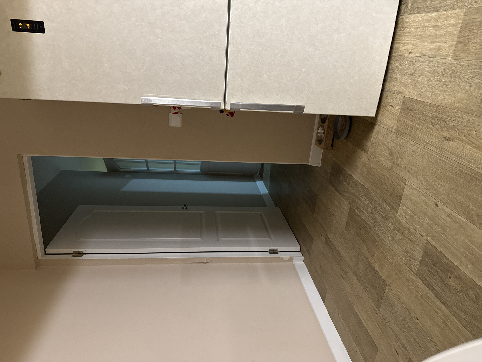 The image shows an interior view featuring a light-colored laminate floor and a wall painted in a light beige. A partially open door leads into another room, with a window visible in the distance. Above the doorway, there is a set of cabinets or a refrigerator with a light-colored finish and silver handles. A pet food bowl is also visible near the edge of the floor. The location is indoors and unnamed, within the city of Unknown, Unknown.