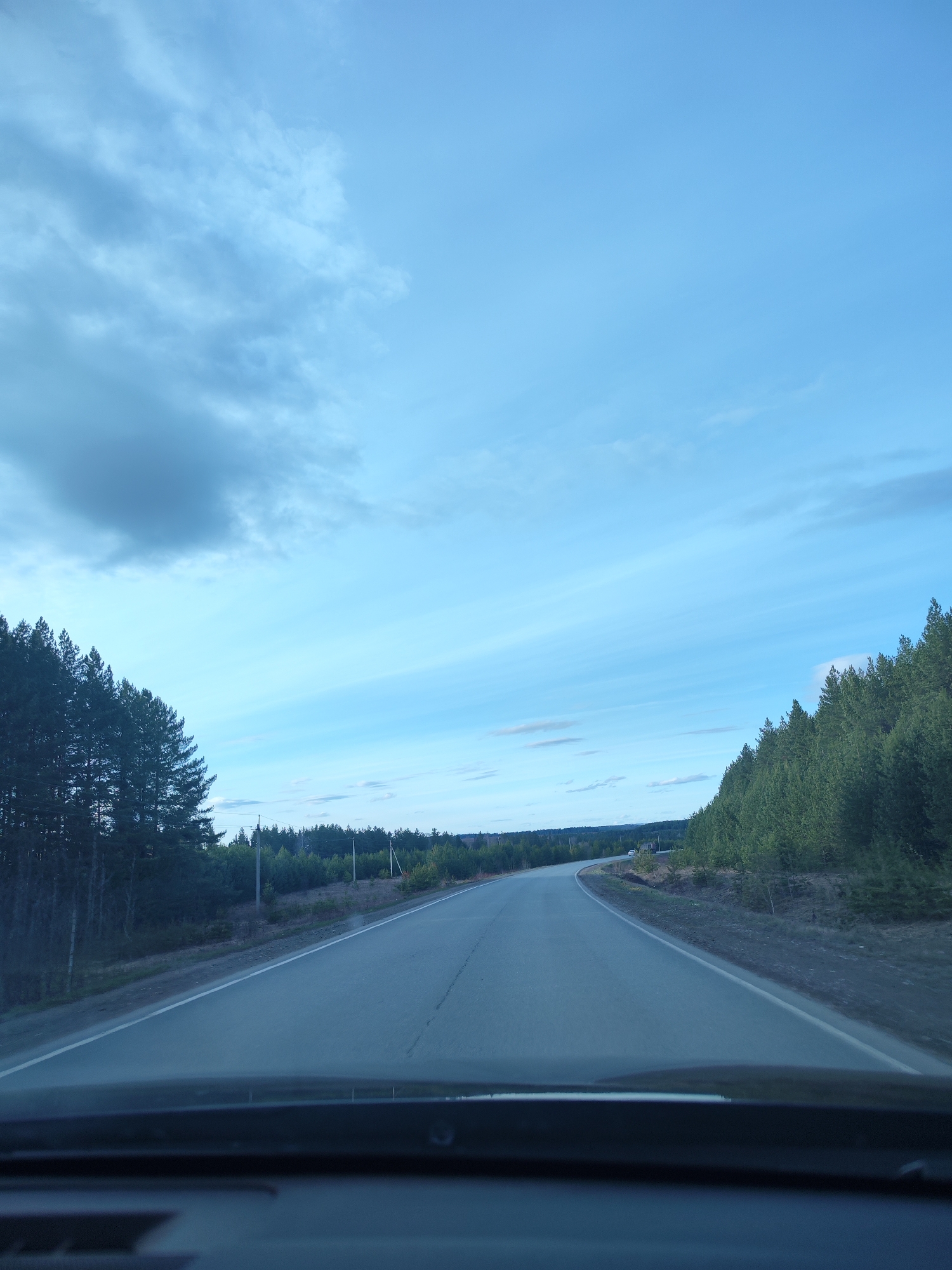 A photograph depicts a paved road stretching into the distance, flanked by dense forests on both sides. The sky above is predominantly blue, with some clouds scattered across the upper left portion of the frame. The perspective suggests the photo was taken from inside a vehicle, as part of the dashboard is visible at the bottom. The location is Unknown, Unknown.