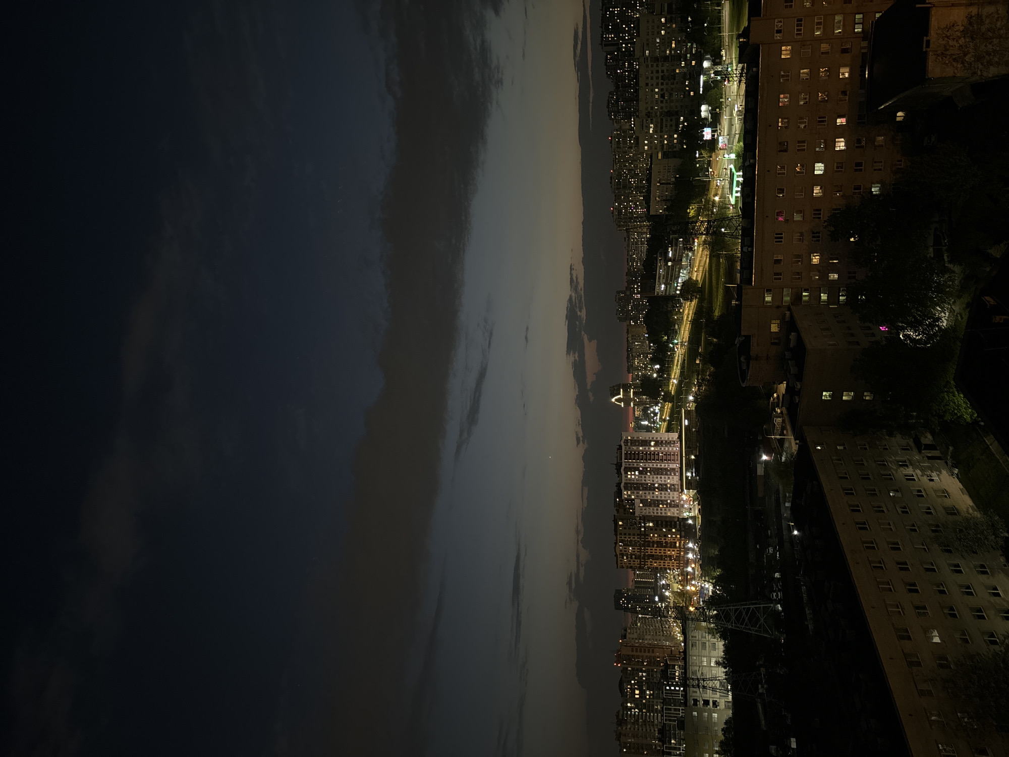 A nighttime aerial view in the city of Unknown, Unknown. On the right is a tall building with many lit windows. The foreground also shows trees and street lights. To the left of the buildings, there is a road with lights illuminating the path. The sky is dark with clouds and some light coming through.