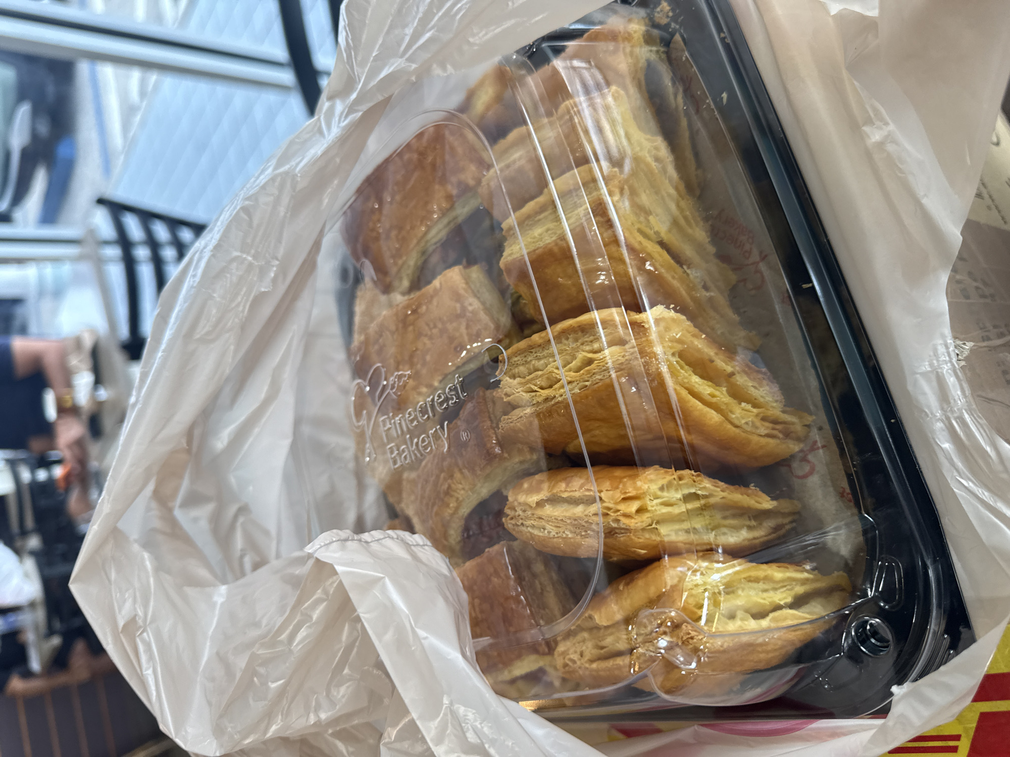 The image shows a clear plastic container filled with pastries from Pinecrest Bakery, inside a white plastic bag. The container has a black rim. The pastries appear to be flaky and golden brown. The background is blurred, showing what seems to be a window with a view of an interior space, possibly a store or cafe. Given the presence of Pinecrest Bakery, it can be inferred that the location is likely in Miami Beach, United States of America, where this bakery has multiple locations.