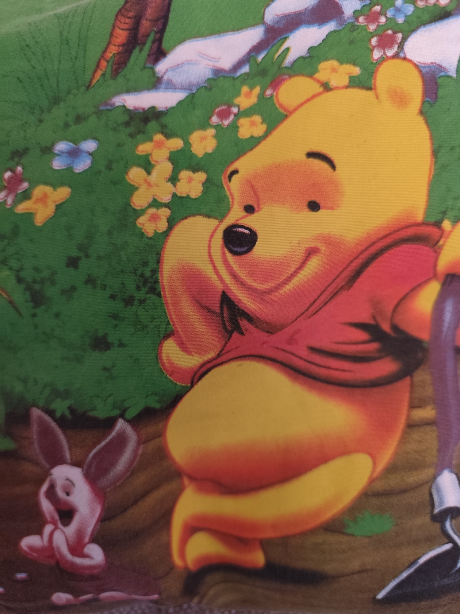 This is a digitally rendered illustration of Winnie the Pooh and Piglet in a garden setting. Winnie the Pooh, a yellow anthropomorphic bear, is depicted from the chest down, wearing a red shirt. He appears to be sitting with his legs crossed, holding a purple gardening tool. Piglet, a small pink pig, is shown to the left, seated with his hands clasped and his mouth open in what appears to be a joyful expression. The background consists of green grass, yellow and pink flowers, and a brown tree trunk. The scene is likely a still from a children's animation or a printed depiction on merchandise. This image has no relation to Montana, Bulgaria, as it is a fictional scene.