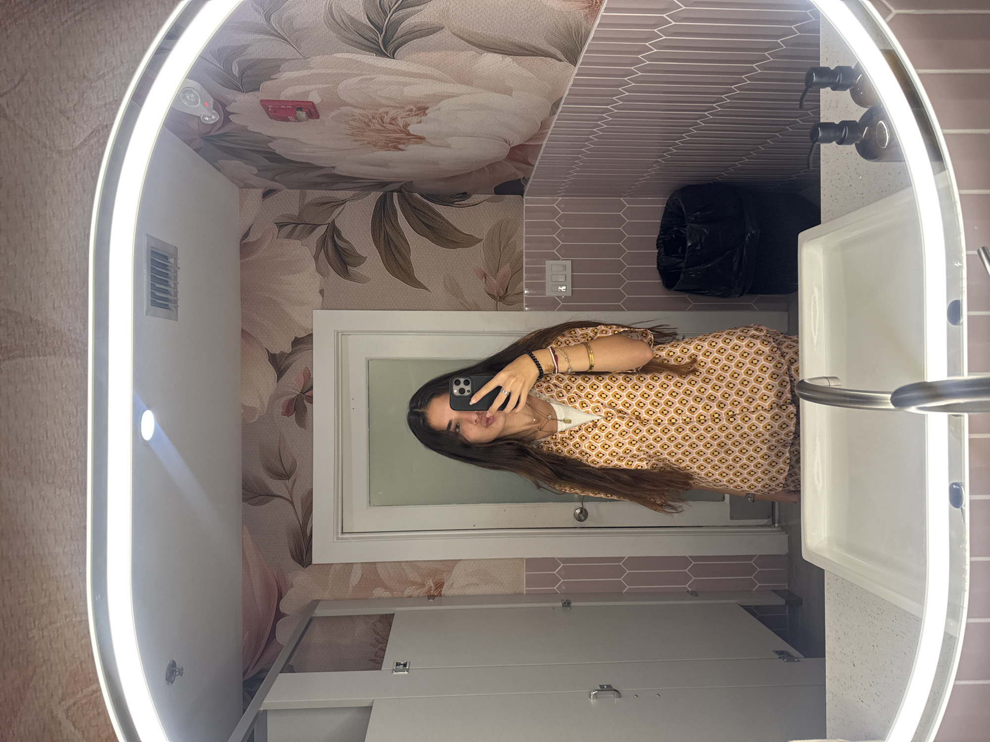 A young woman with long dark hair takes a mirror selfie in a well-lit bathroom. She is wearing a short-sleeved collared shirt with an orange and yellow geometric pattern and a white undershirt. She is also wearing several bracelets and necklaces. She is holding a black smartphone in her right hand, with which she is taking the picture. The bathroom has floral wallpaper with large pink and white flowers. There are two stalls with white doors on the left side of the image. A white sink with a silver faucet is in the foreground. There is a black trash can behind the sink and a red fire alarm on the right wall. The bathroom is located in Boca Raton, United States of America.