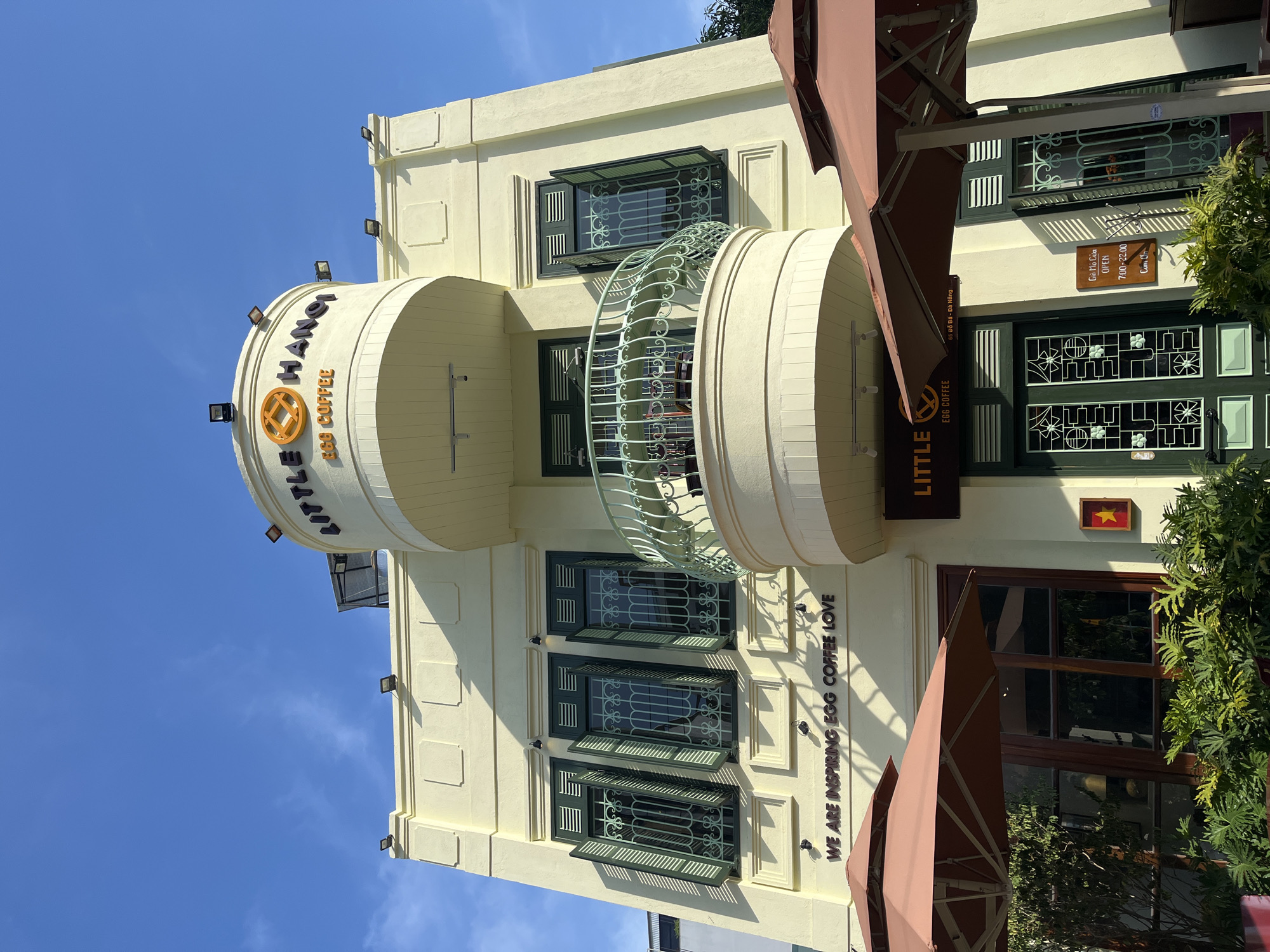 The image displays the exterior of a light yellow building, identified as a cafe named "LITTLE HANOI EGG COFFEE," under a clear blue sky in Đà Nẵng, Vietnam. The building features several windows with dark green frames and decorative grilles, some having open shutters. A prominent arched balcony with an ornate light green railing is situated on the second level.
Above the balcony, a large circular sign spells out "LITTLE HANOI EGG COFFEE" in dark letters with an orange logo. Below this, on the building's wall, white text declares "WE ARE INSPIRING EGG COFFEE LOVE." On the right side, partially obscured by extended brown awnings, another vertical sign reads "LITTLE EGG COFFEE." Near the entrance, a wooden sign provides key information: "65 Đỗ Bá - Đà Nẵng" (the address), "Giờ Mỏ Cửa (Opening Hours) OPEN 7:00-22:00", and "Cám Ơn" (Thank You). A small framed image resembling the Vietnamese flag (yellow star on a red background) is also visible.
The scene is brightly lit by the sun, indicating clear weather, likely mid-morning or early afternoon, with shadows cast on the building's facade. Several small spotlights are mounted on the exterior walls. Green foliage from potted plants or bushes adds natural elements to the bottom right of the frame, contributing to a welcoming ambiance for the establishment.