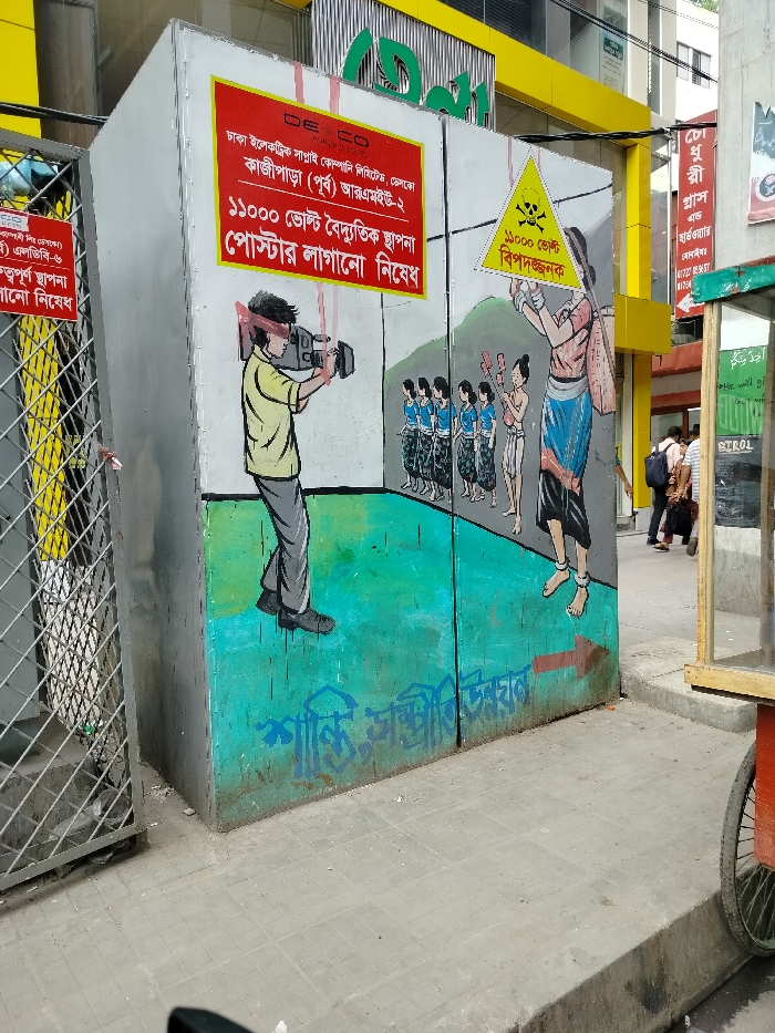 The image shows a street scene in Dhaka, Bangladesh, at the coordinates 23.763° N, 90.39° E. A large, gray, rectangular electrical box sits on the sidewalk. The side facing the viewer is covered with a colorful mural. The left half of the mural is a yellow and green illustration that depicts a man wearing a yellow shirt and blue pants, holding a large camera in his hands, filming a group of people. The right half of the mural is a green and blue illustration depicting a woman in a blue and white dress, holding a red flag, and walking on the street. The scene is crowded with people in the street, both standing and walking, as well as signs, streetlights and buildings with yellow accents. A red arrow on the mural points to the right. The sidewalk is made of gray concrete. There is a wooden stand with a metal rack on the right side of the scene. The stand is holding a glass case with goods.