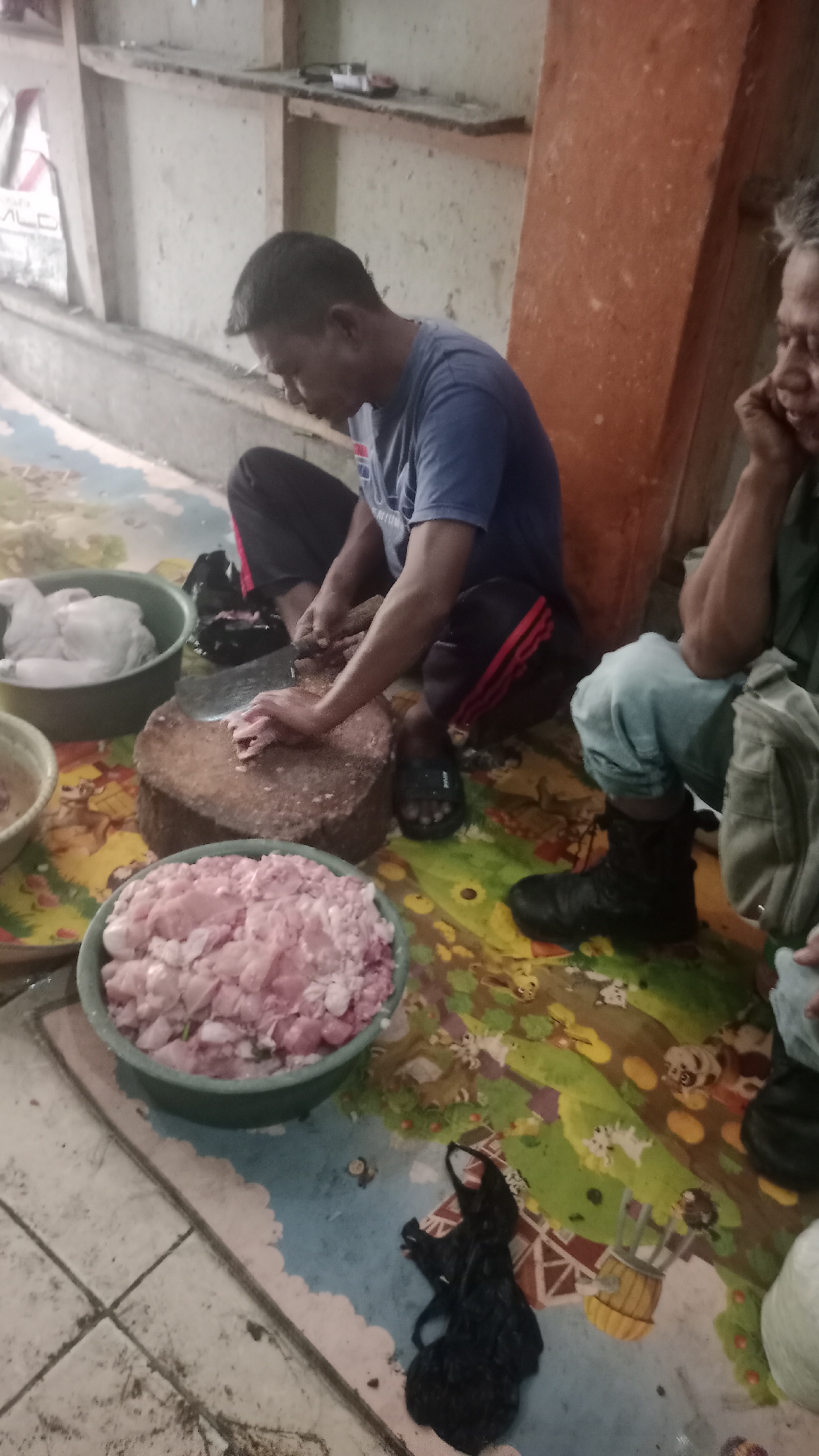 In Karyamandala, Indonesia, two individuals are engaged in food preparation outdoors. A man in a blue t-shirt and black pants with red stripes is crouched on the ground, using a large cleaver to chop meat on a thick wooden cutting board. In front of him is a green bowl filled with chopped pink meat. To his left, another green bowl contains white, possibly raw, chicken. To his right, another person, wearing light blue pants and a dark boot, is also seated on the ground. They appear to be talking on a phone and have a light-colored bag to their right. The ground is covered with a colorful, patterned mat depicting cartoon animals. In the background, a rough, light-colored wall and a wooden shelf with some items are visible.