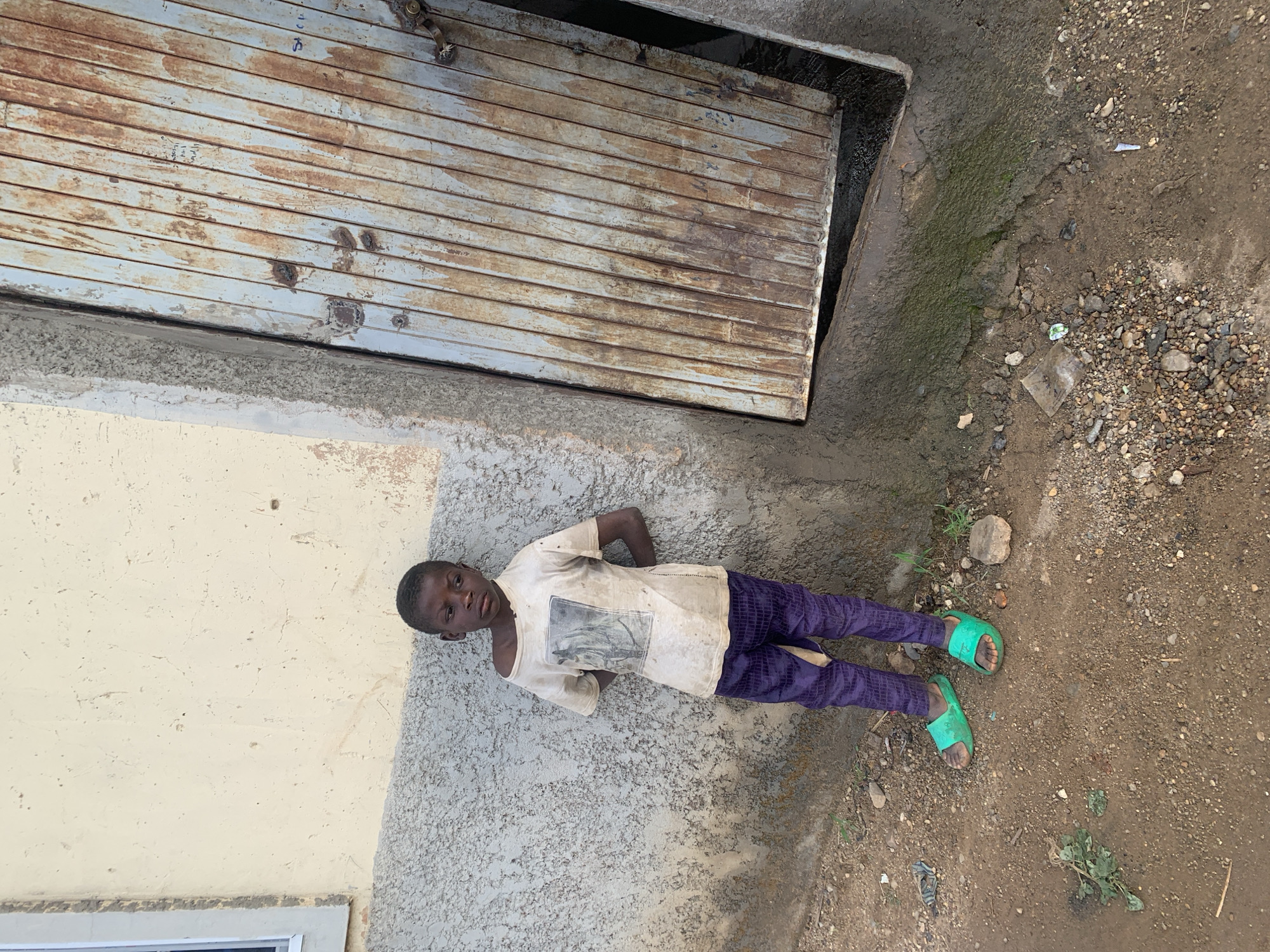 A young person lies on the ground, adjacent to a building. The individual is wearing a light-colored t-shirt, dark purple pants, and green sandals. The building features a rolling metal door and tan-colored walls. The ground is dirt and gravel. The location is Jos, Nigeria.