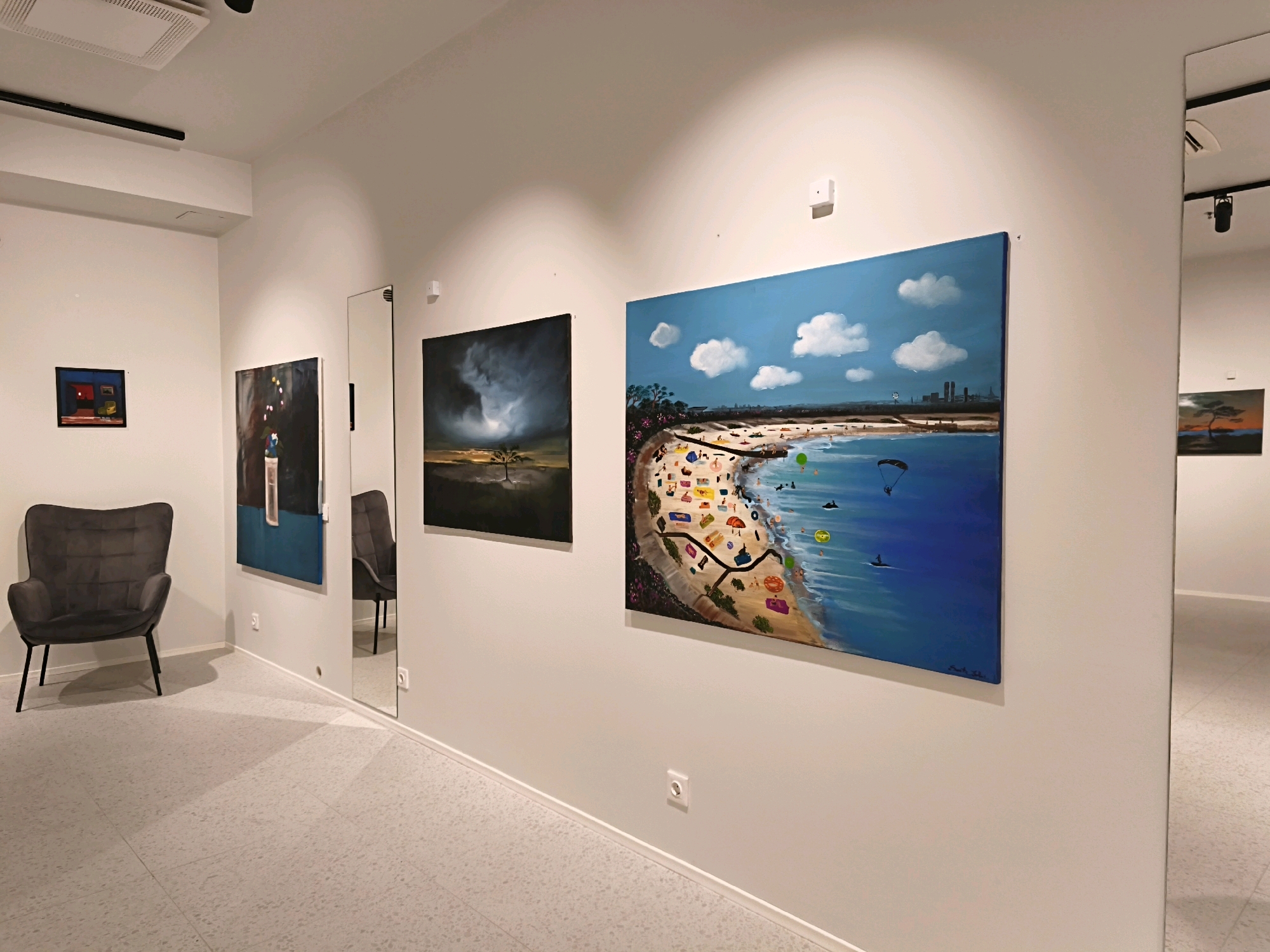 The image depicts the interior of a contemporary art gallery. The walls are painted white, and the floor is a light-colored speckled material. Several artworks are displayed on the walls. In the foreground on the left, a dark gray armchair with a tufted back and black legs is placed. To the right of the armchair, a large vertical mirror leans against the wall.
On the wall to the left of the mirror, there is a small framed painting with a dark background, a red object, and a yellow object. Next to it, a larger vertical painting features abstract shapes with a dominant blue element and a vertical black and white section. To the right of the mirror, a large horizontal painting shows a dramatic dark sky with a single tree in a desolate landscape. Further to the right, a vibrant, large horizontal painting depicts a crowded beach scene with numerous colorful umbrellas, people, and various water activities under a bright blue sky with white clouds. A cityscape is visible in the distance.
The lighting in the gallery appears to be track lighting mounted on the ceiling, casting shadows and highlighting the artwork. The environment suggests a well-lit and modern exhibition space. There are no people visible in the image. The specific location in Tallinn, Estonia, cannot be determined from the visual information provided.