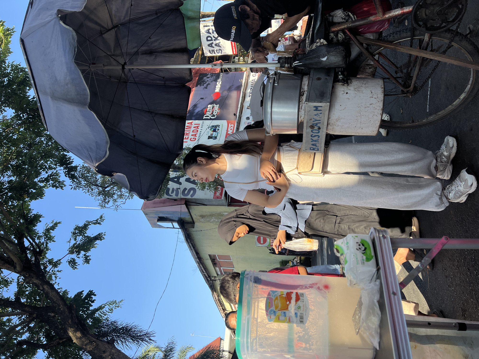 In Kecamatan Pati, Indonesia, a woman in a white crop top and grey sweatpants stands near a food cart. The food cart, partially covered by a black umbrella, displays a sign reading "BAKSO AYAM." A man wearing a black cap is operating the cart. In the background, buildings with colorful advertisements are visible. Other people are present, including a woman in a black hijab. A large plastic container is on a table in the foreground. The scene appears to be a street market or food stall area under a clear blue sky.