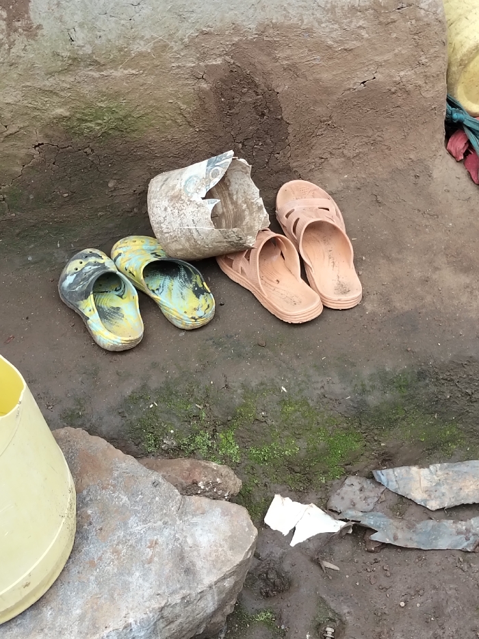 The scene depicts an outdoor ground-level area in Nyansiongo ward, Kenya. The ground is composed of dark, damp soil with patches of green moss. A cracked, textured earthen or plastered wall serves as the background. In the center, two pairs of worn plastic footwear are visible: one pair of mottled yellow/green and black clogs, and one pair of light brown open-toed sandals. Between the footwear lies a severely broken, light-colored plastic container. In the lower-left quadrant, a portion of a yellow plastic bucket rests on a large gray rock. Scattered debris, including dark fragments (possibly metal or plastic) and a piece of white paper, are present on the ground in the lower-right.