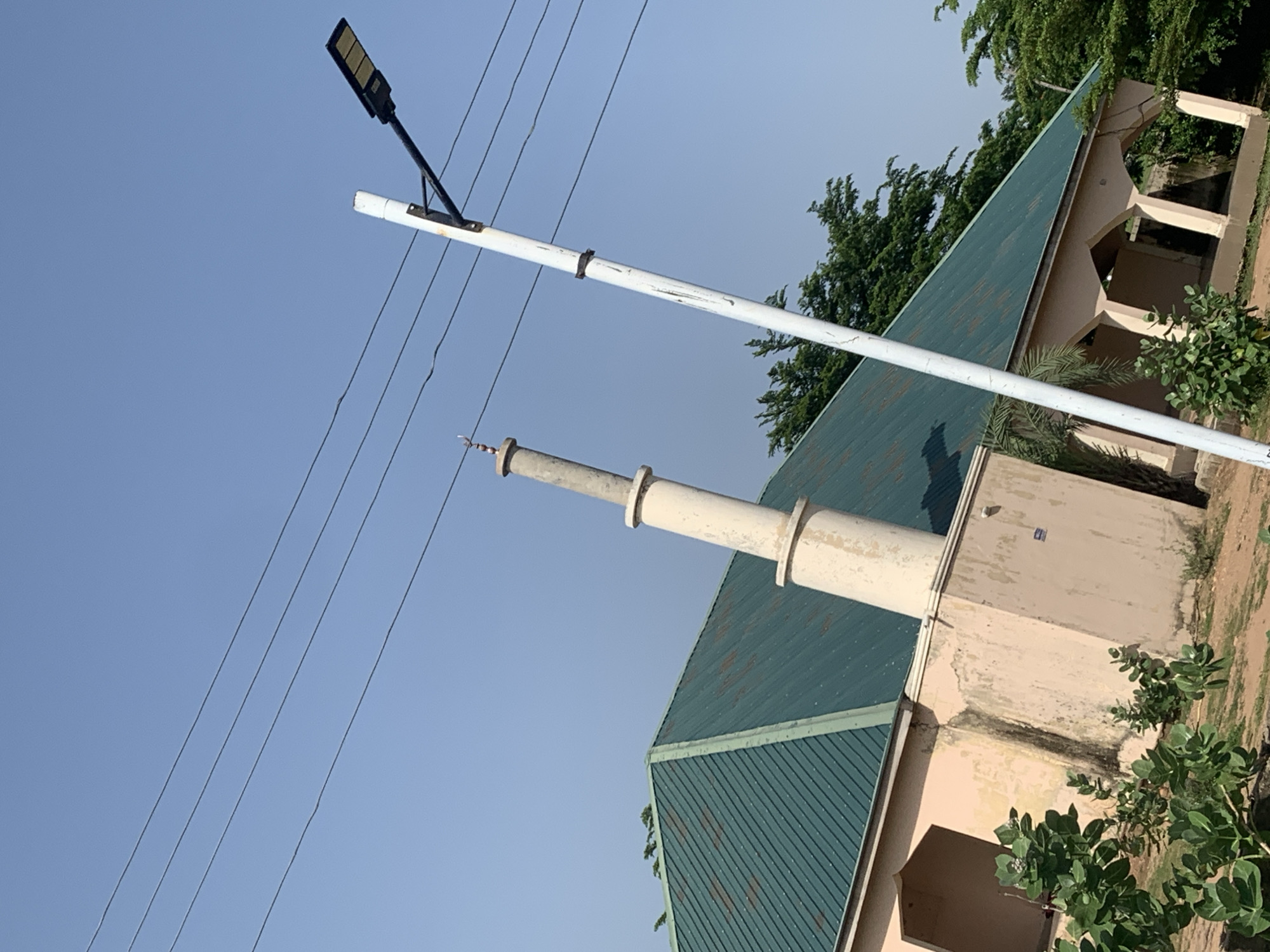 The image captures an exterior view of a building in Jalingo, Nigeria, under a clear blue sky. A prominent green, corrugated metal roof covers a portion of the structure, which has light beige, stuccoed walls. To the left of the building, a tall, slender, cream-colored minaret with rounded bands rises towards the sky. A white utility pole with a black modern street lamp attached is positioned in the foreground, slightly to the right of the minaret. Several black power lines are strung across the upper portion of the frame. Lush green foliage, including some bushes and trees, surrounds the building and extends into the background. The overall scene suggests a daytime setting in a semi-urban or rural environment.