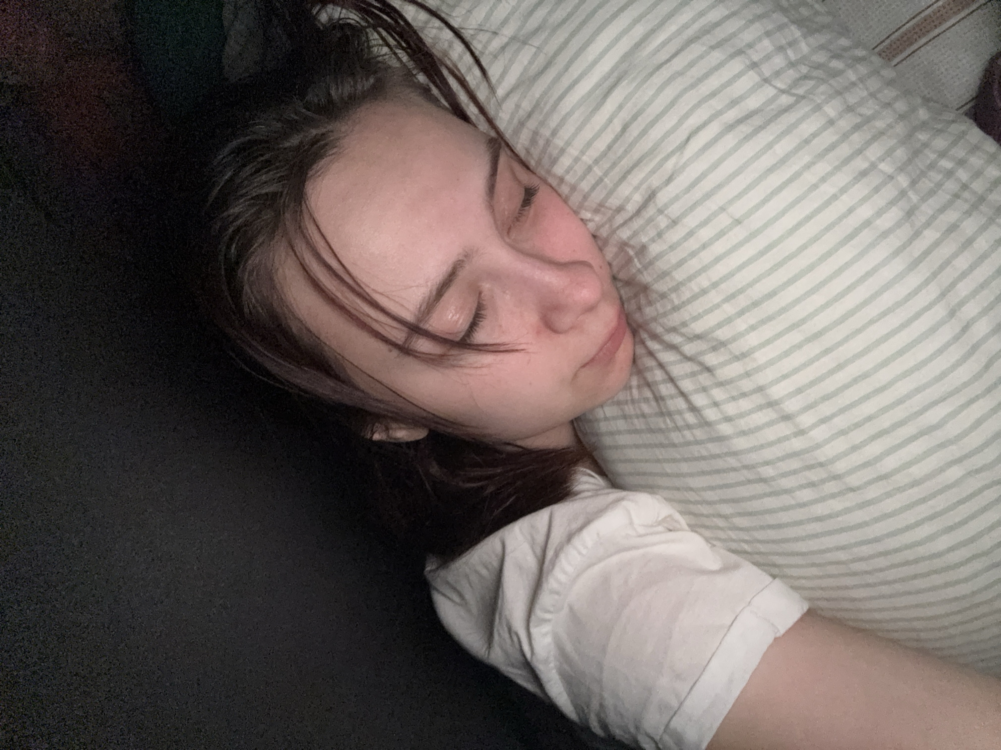 A young woman is asleep on a bed. She is lying on her side, her head resting on a striped pillow. She is wearing a white shirt. The sheets are black.
I cannot determine the location of the photo.