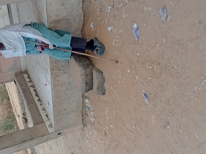 A person is captured in a sideways, mid-shot. Dressed in a white shirt and blue shorts, they hold a brown stick. The setting involves a corner of a building with visible steps and a concrete base. The ground is dirt, scattered with bits of trash, appearing earthy and dry.