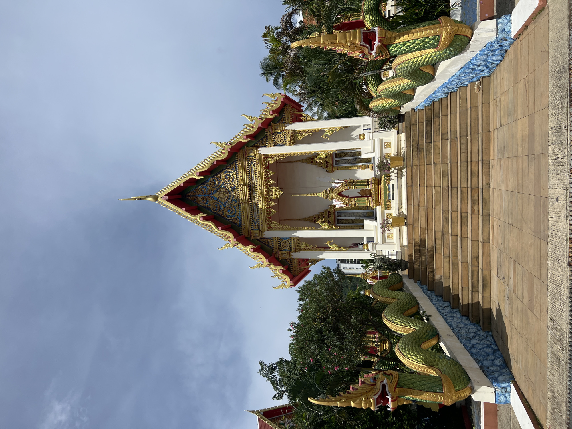 A vibrant Buddhist temple with ornate golden decorations sits at the top of a wide, tiled staircase in Karon, Thailand. The temple features a steeply pitched roof with intricate patterns and red and gold accents, topped by a spire. Flanking the staircase are two large, green and gold serpent statues, each with a fearsome head and a scaled body that winds along the railings. The temple building is white with gold trim and windows, and at its base are smaller decorative structures and potted plants. Lush green trees and palm fronds surround the temple, and a cloudy blue sky with a few wisps of white clouds forms the background. The overall scene is one of religious grandeur and tropical lushness.