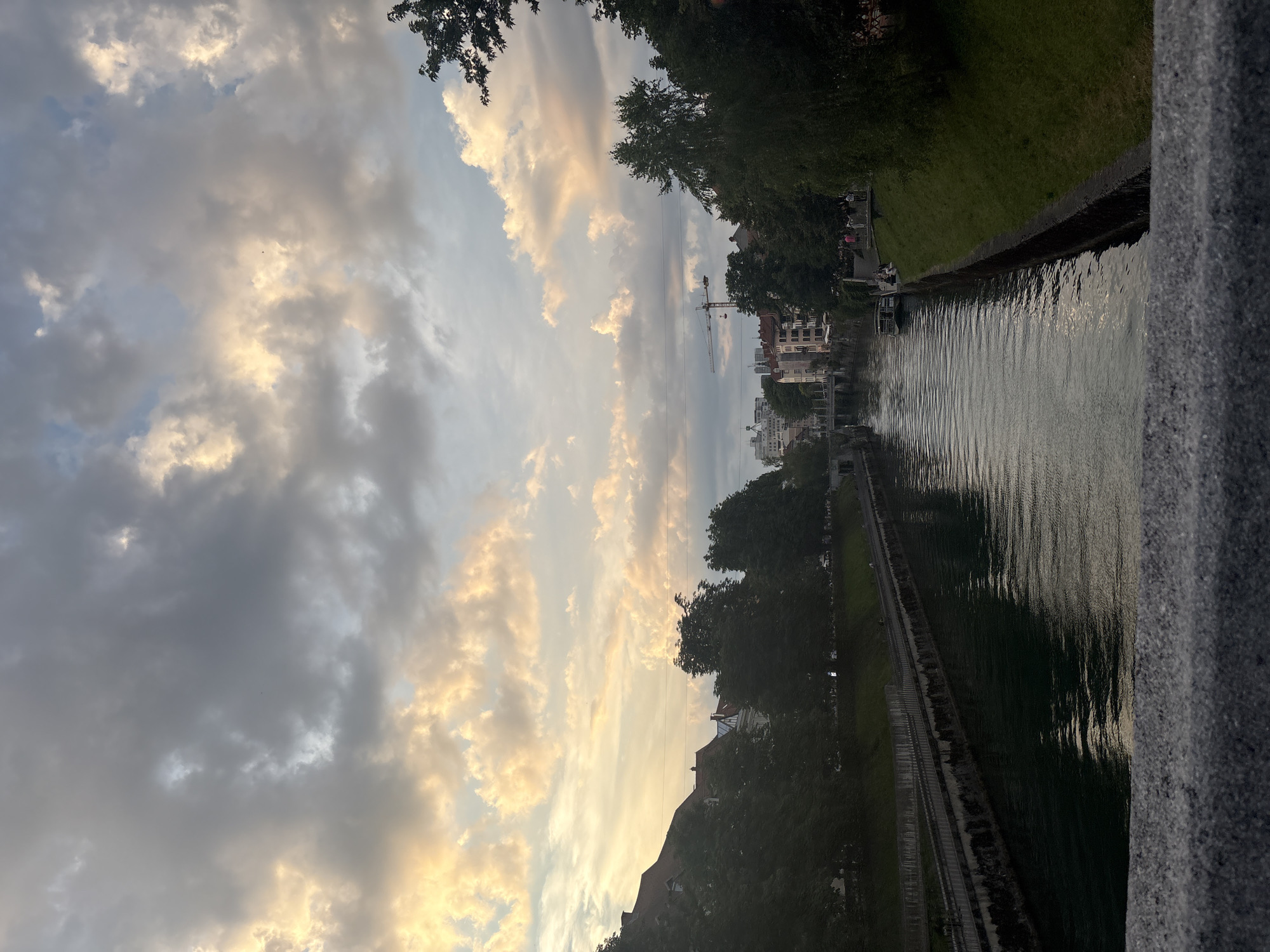 The image captures a view of a river canal in a city during what appears to be sunset or late afternoon. The sky is filled with dramatic, textured clouds, some illuminated by warm, golden light, while others are a muted gray. On the left bank of the canal, there are buildings with red-tiled roofs, interspersed with trees. On the right bank, a grassy slope lined with stone borders the canal, and further back, more trees and some buildings are visible. A small boat is moored on the right side of the canal. In the distance, a construction crane stands against the sky, suggesting ongoing development. The water of the canal is dark and reflects the muted colors of the sky. In the foreground, the edge of a stone railing or bridge is visible, out of focus. A few people can be seen walking or sitting along the canal banks. The scene suggests a peaceful urban environment by the water.