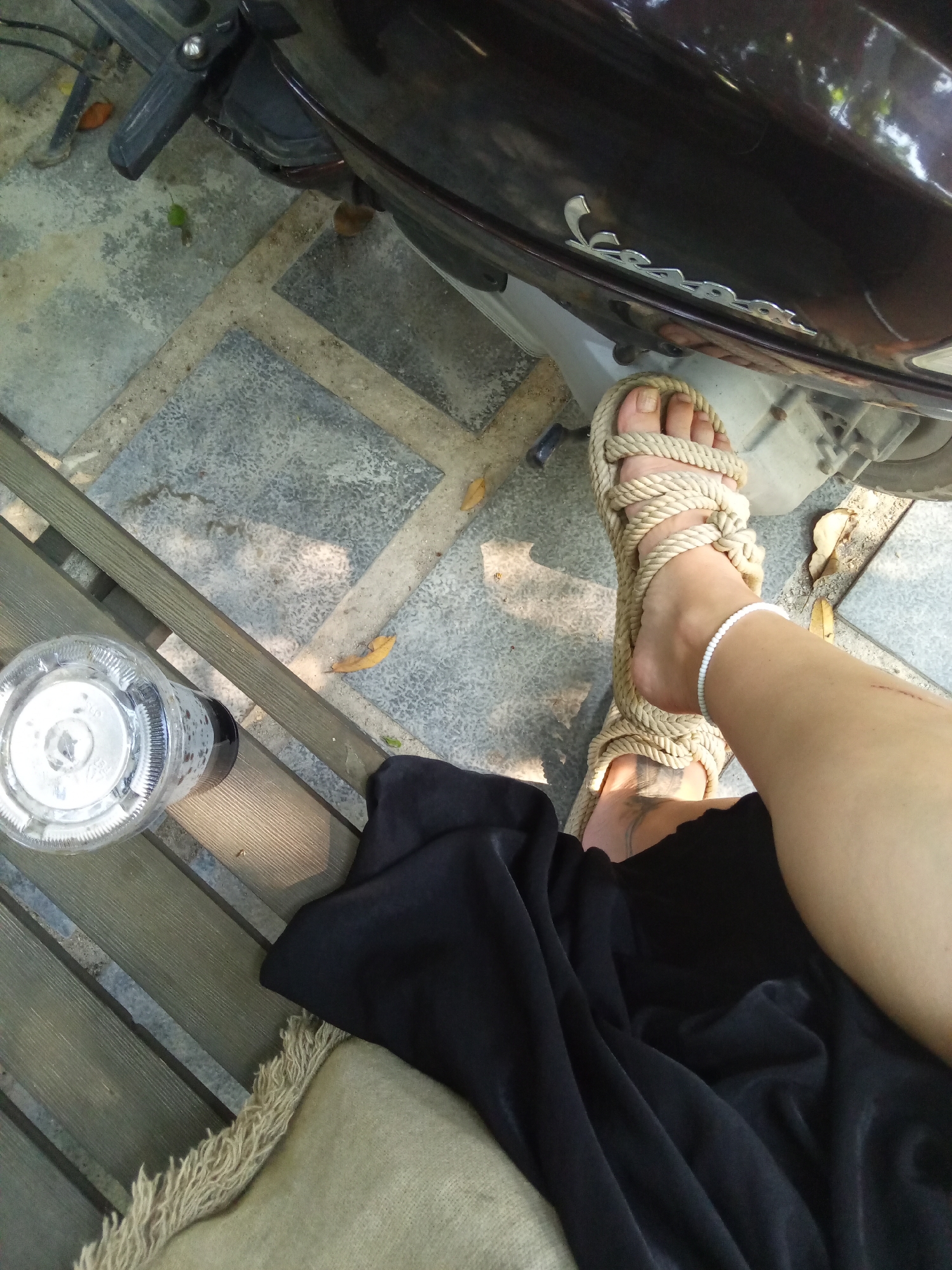 A partially visible person sits on a wooden bench next to a brown Vespa scooter parked on a tiled surface. The person wears a black garment and a pair of beige rope sandals, adorned with a white beaded anklet. A clear plastic cup with a dark liquid is on the bench. The background is the city of Bac Natural Heritage Area Trang, Vietnam.