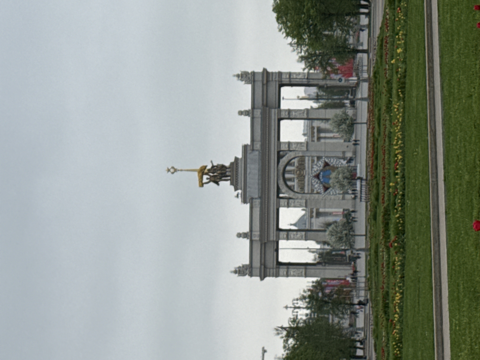 The image shows the Central Pavilion at the Exhibition of Achievements of National Economy (VDNKh) in Moscow, Russia. The pavilion is a large, grey structure with a golden statue on top depicting a worker and a collective farm woman holding a sheaf of wheat. Arches are visible in the pavilion's design. In the foreground is a flowerbed with red and yellow flowers, alongside a strip of green grass. The sky is overcast.