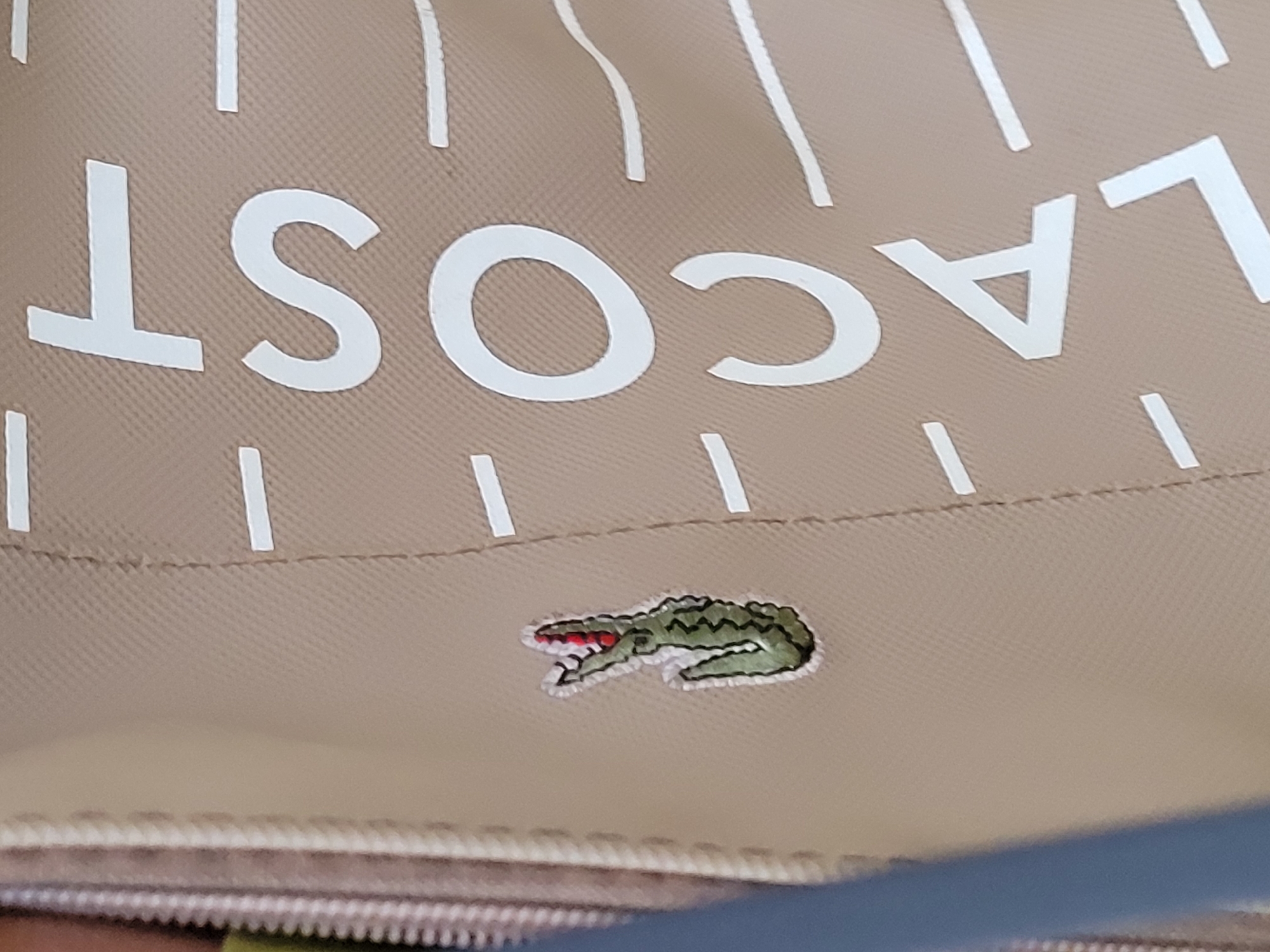 The image shows a close-up of a beige bag with the word "LACOST" printed in large white letters. Below the word is a small embroidered green crocodile logo with a red mouth. The fabric of the bag appears to be textured. The image does not contain any information relating to Gura Top, Nigeria.