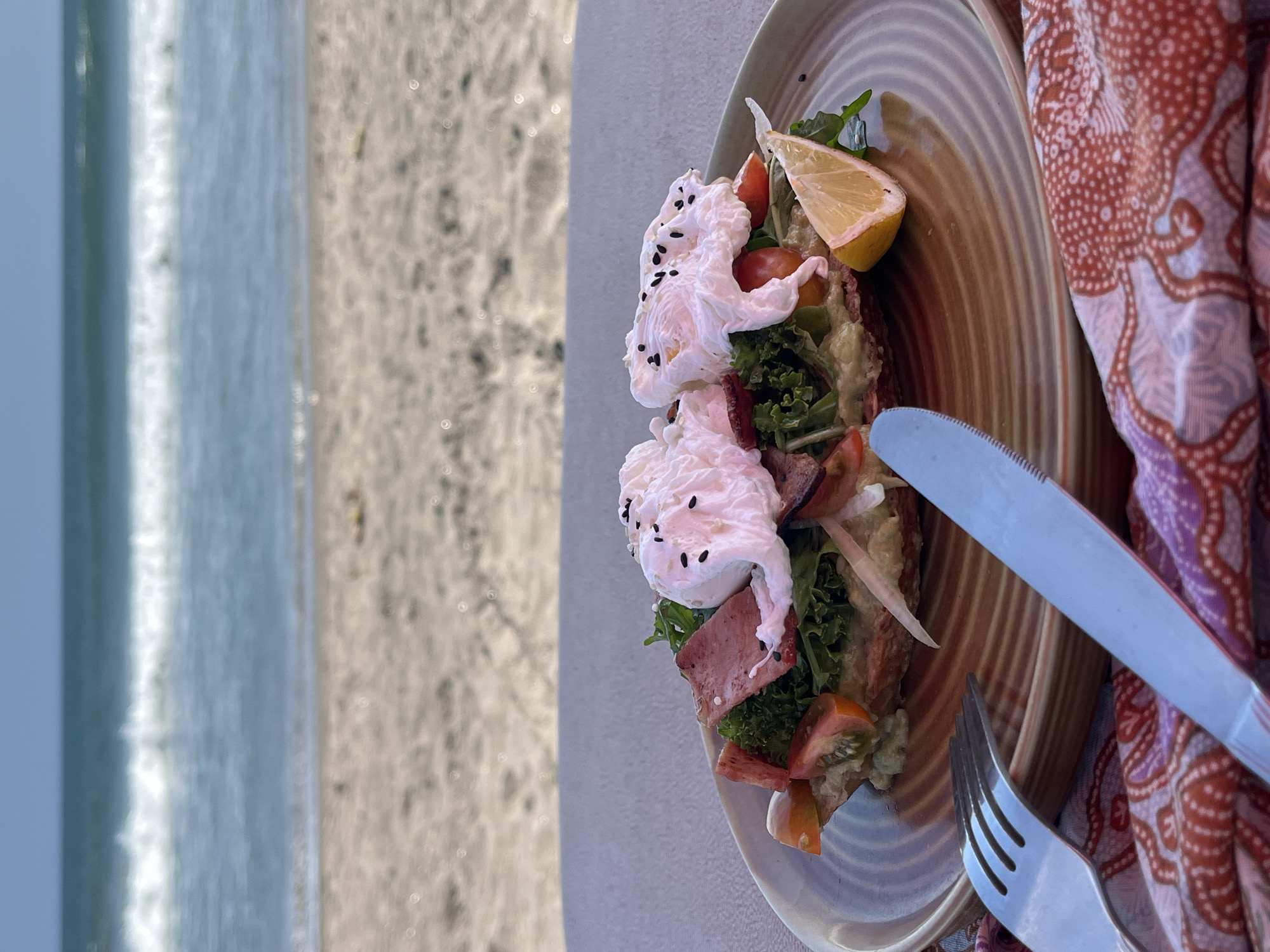 A close-up, eye-level shot showcases a savory open-faced sandwich on a brown, circular plate with a textured, ribbed surface. The sandwich is generously topped with two poached eggs, their whites delicately draped over a bed of greens, thinly sliced tomatoes, and what appears to be ham or cured meat. Black sesame seeds are sprinkled over the eggs, adding a visual contrast. A wedge of lemon sits on the edge of the plate.
The plate rests on a taupe-colored surface, likely a table, set against a backdrop of a beach scene. In the background, the sandy shore meets the ocean, with gentle waves breaking on the beach under a clear, pale blue sky.
To the left of the plate, a fork and knife are positioned, with the fork in the foreground and the knife slightly behind it. Both are made of polished metal. A patterned fabric, predominantly in shades of red and purple with a repeating organic motif, is folded beneath the cutlery and plate, partially obscuring the table's edge.
The scene is set outdoors, suggesting a meal enjoyed with a view of the sea. The lighting indicates it is daytime, likely late afternoon or early morning due to the soft quality of the light. The image is sharp and focused on the food and the immediate setting, with the background slightly softened. The location is identified as North Kuta, Indonesia.