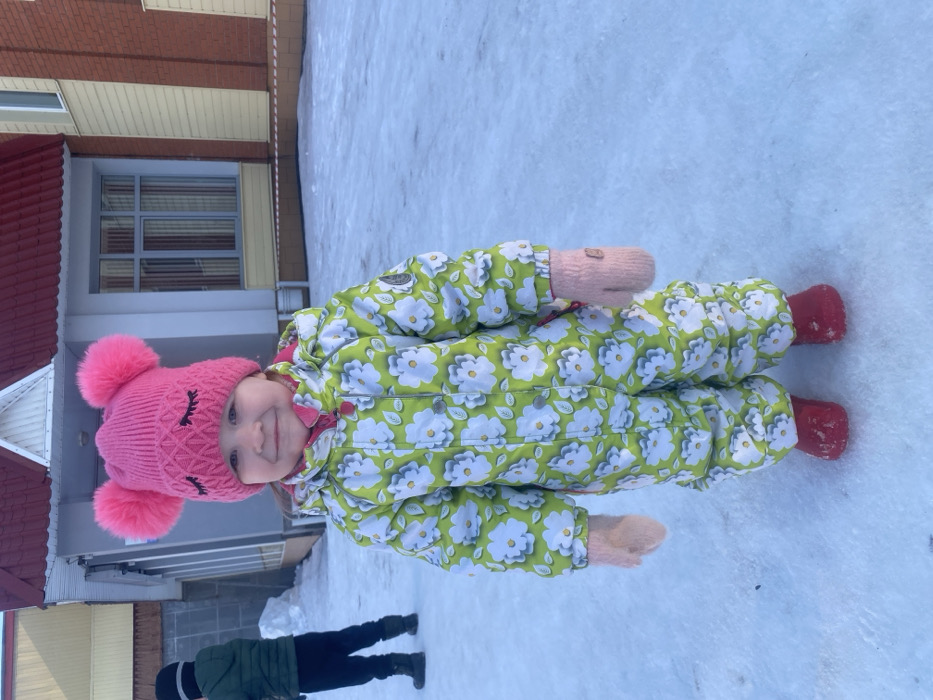 An outdoor scene in Chaikovsky, Russia, depicts two children on snow-covered ground in front of a building. In the foreground, a young child, appearing female, stands centrally, facing the camera with a slight smile. She is dressed in a pink knitted hat with two large pink pom-poms and an embroidered closed-eye design, a lime-green one-piece snowsuit patterned with white flowers and green leaves, pink knitted mittens, and red boots. To the left in the background, another child in a dark green hooded jacket, dark pants, and black boots is partially visible, facing away from the camera. The ground is uniformly covered in snow. Behind the children, a building structure is visible, featuring a section with a light gray facade, a dark red sloped roof with a white triangular gable, and a window with vertical blinds. Adjacent to this, another section of the building displays red brick siding over a cream-colored lower panel.