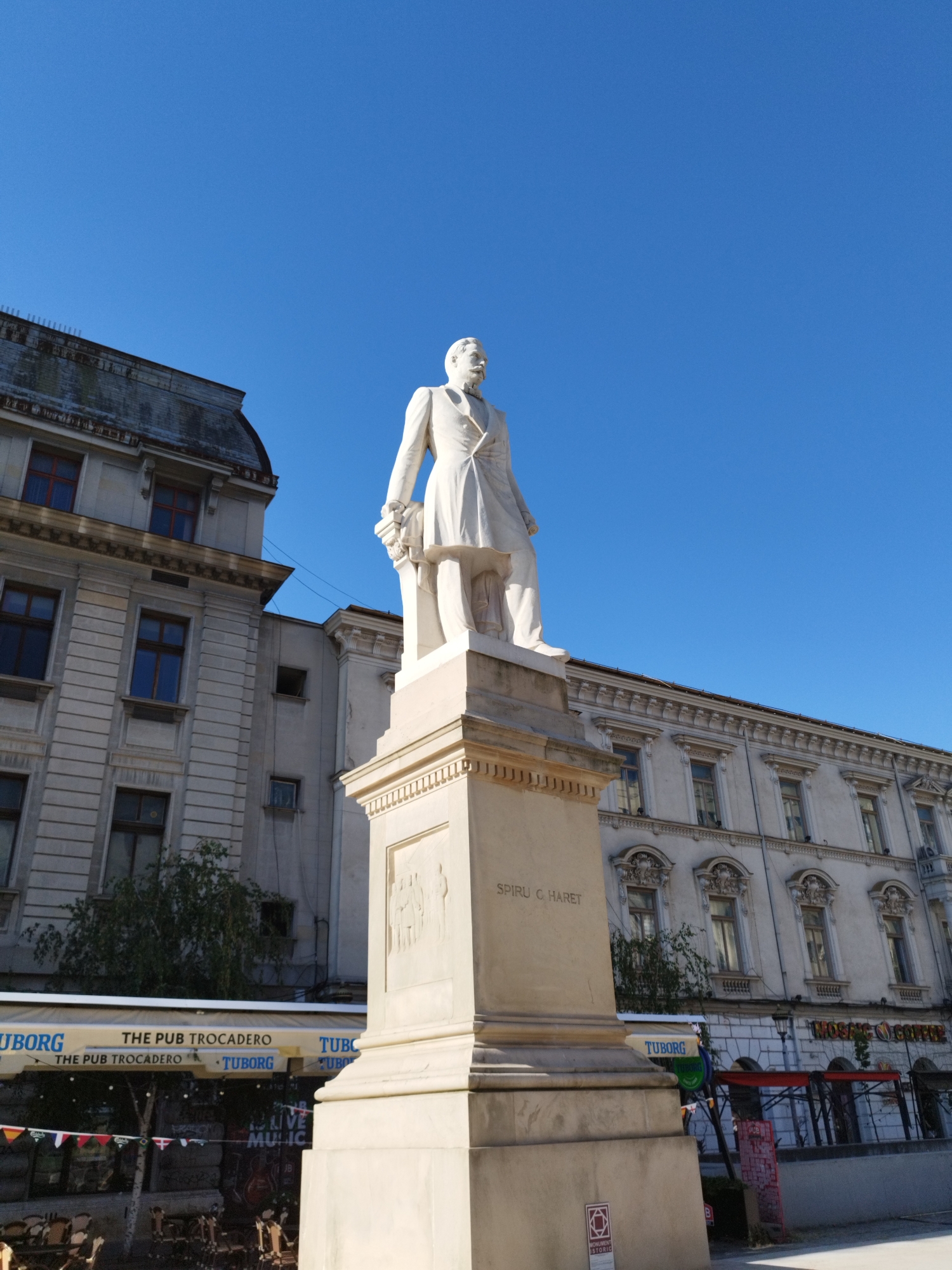 The photograph captures a statue of Spiru C. Haret in Bucharest, Romania, under a clear blue sky. The statue, made of light-colored stone, depicts a man in a formal suit holding a bouquet of flowers. It stands on a tall, ornate pedestal that has inscriptions and relief carvings on its sides. To the left of the statue, a large, old building with multiple windows and a grey, sloped roof is visible. To the right, another similar building with many windows and decorative elements is partially seen. In the foreground, a row of awnings for a establishment named "THE PUB TROCADERO" is present, with some tables and chairs set up outside. The area appears to be a public space with architectural significance.