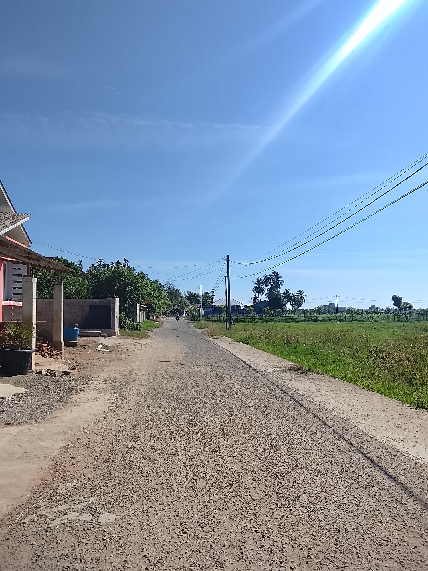 The image depicts a rural road in Geuleudah, Indonesia, under a clear blue sky. The road is paved with gravel and leads into the distance, flanked by green fields on the right and a residential area on the left. Several power lines are visible overhead, stretching across the frame. On the left side of the road, a house with a pink facade and a covered porch is partially visible, along with some construction materials. Further down the road, a motorcycle can be seen in motion. The overall scene suggests a peaceful, sunny day in a semi-urban or rural setting.