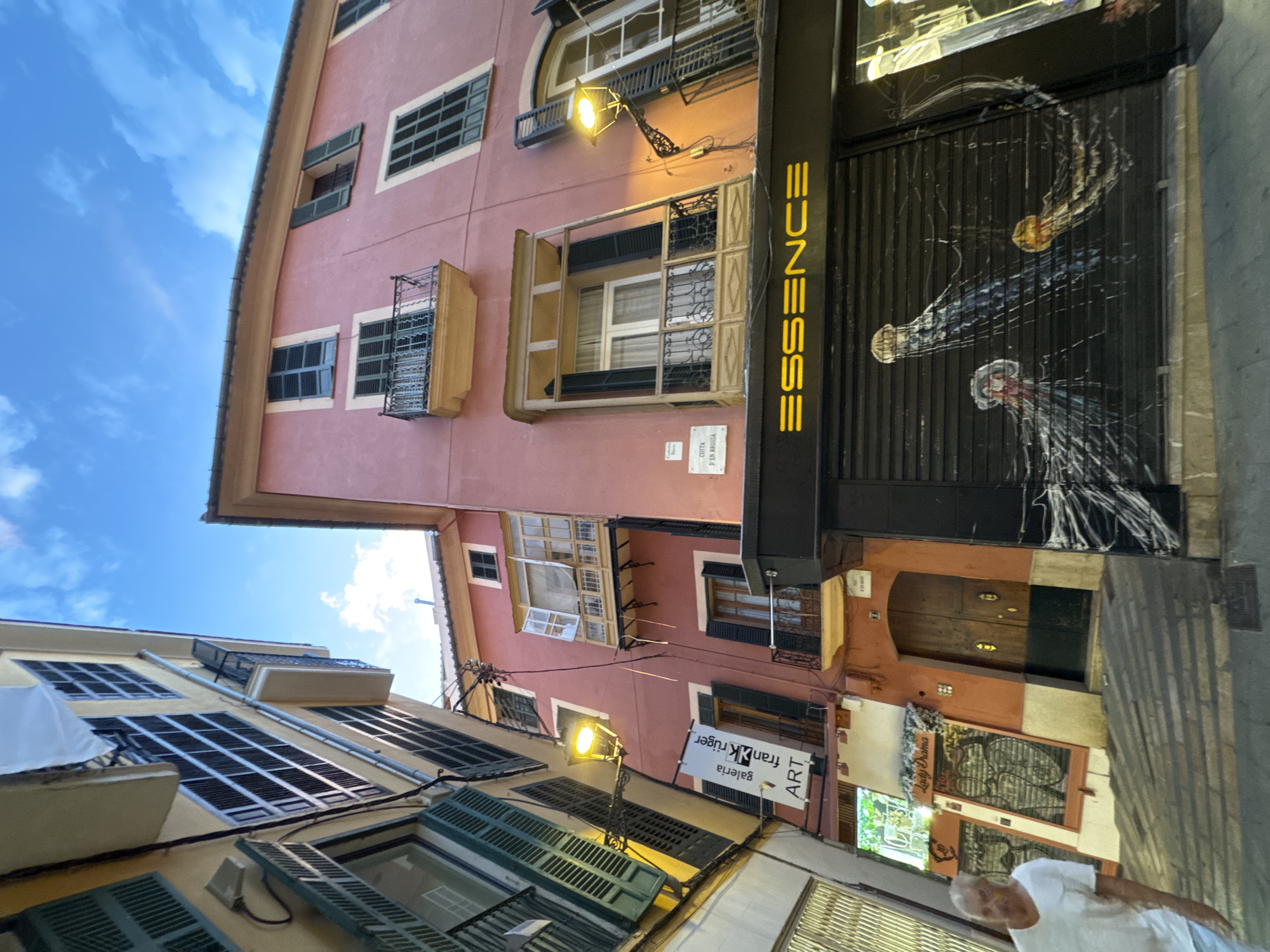This is a street-level, wide-angle photograph taken in Palma de Mallorca, Spain, looking up at buildings lining a narrow street. The buildings are a mix of architectural styles and colors, with the pink building dominating the right side of the frame and a yellow building on the left. The sky is blue with scattered clouds.
On the pink building, there are several windows with dark green shutters, some of which are open. A small balcony with black iron railings protrudes from one of the windows. Below this, a black metal shuttered storefront displays the illuminated yellow text "ESSENCE". The shutter itself is decorated with a mural of three jellyfish. Above this storefront, a narrow balcony with a wooden frame surrounds a window. Further up the pink building, another balcony is visible.
The yellow building on the left features numerous windows with dark green shutters, some of which are also open. A yellow streetlamp is attached to the side of this building, casting a warm light. A banner hangs from the yellow building, reading "galeria fran Krüger ART".
A portion of a man wearing a white t-shirt is visible on the left side of the image, walking away from the camera. The street itself is paved with cobblestones, and a few other details, such as a door and some graffiti on a lower level storefront, are also present. The overall impression is of a charming, narrow European street with a mix of residential and commercial spaces.