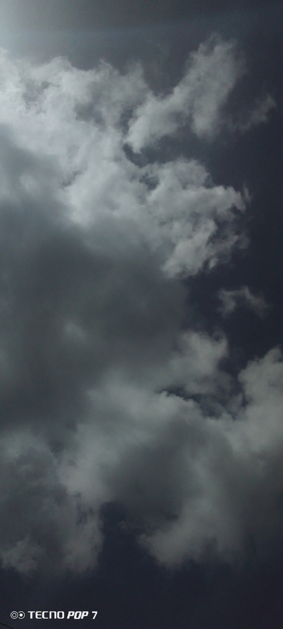 The image shows a sky filled with dark, ominous clouds. The sky appears to be an overcast day or approaching a storm, with little to no blue visible. No people or buildings are visible. The camera model indicated on the bottom left of the image is TECNO POP 7. The location, Altagracia, Venezuela, is inferred.