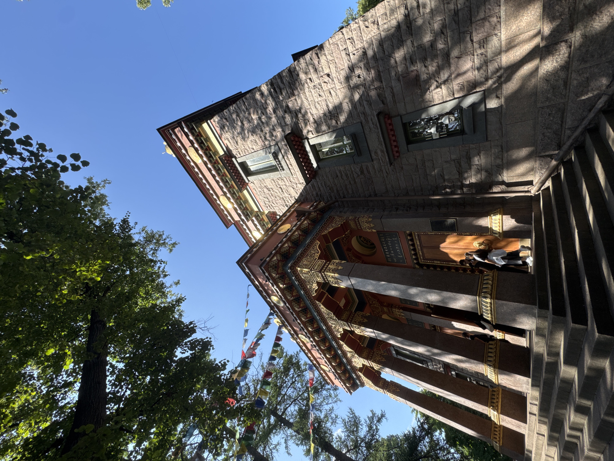 The image shows the Buddhist Temple (Datsan) in Saint Petersburg, Russia, on a sunny day. The camera is positioned low, looking up at the temple's facade. The temple is constructed of large, rough-cut stones and features ornate, colorful architectural elements, including red and gold decorations on the roofline and pillars. Prayer flags in various colors are strung between trees and the temple.
The temple has a prominent entrance with large wooden doors, flanked by several tall, decorative pillars. Two individuals are visible near the entrance, one walking away from the camera and another standing to the side. The steps leading up to the temple are wide and stone. Sunlight casts dappled shadows from the trees onto the temple's stone walls and the steps. The sky is a clear, bright blue. A large tree with dense green foliage is visible in the upper left portion of the image, partially obscuring the sky and the top of the temple.