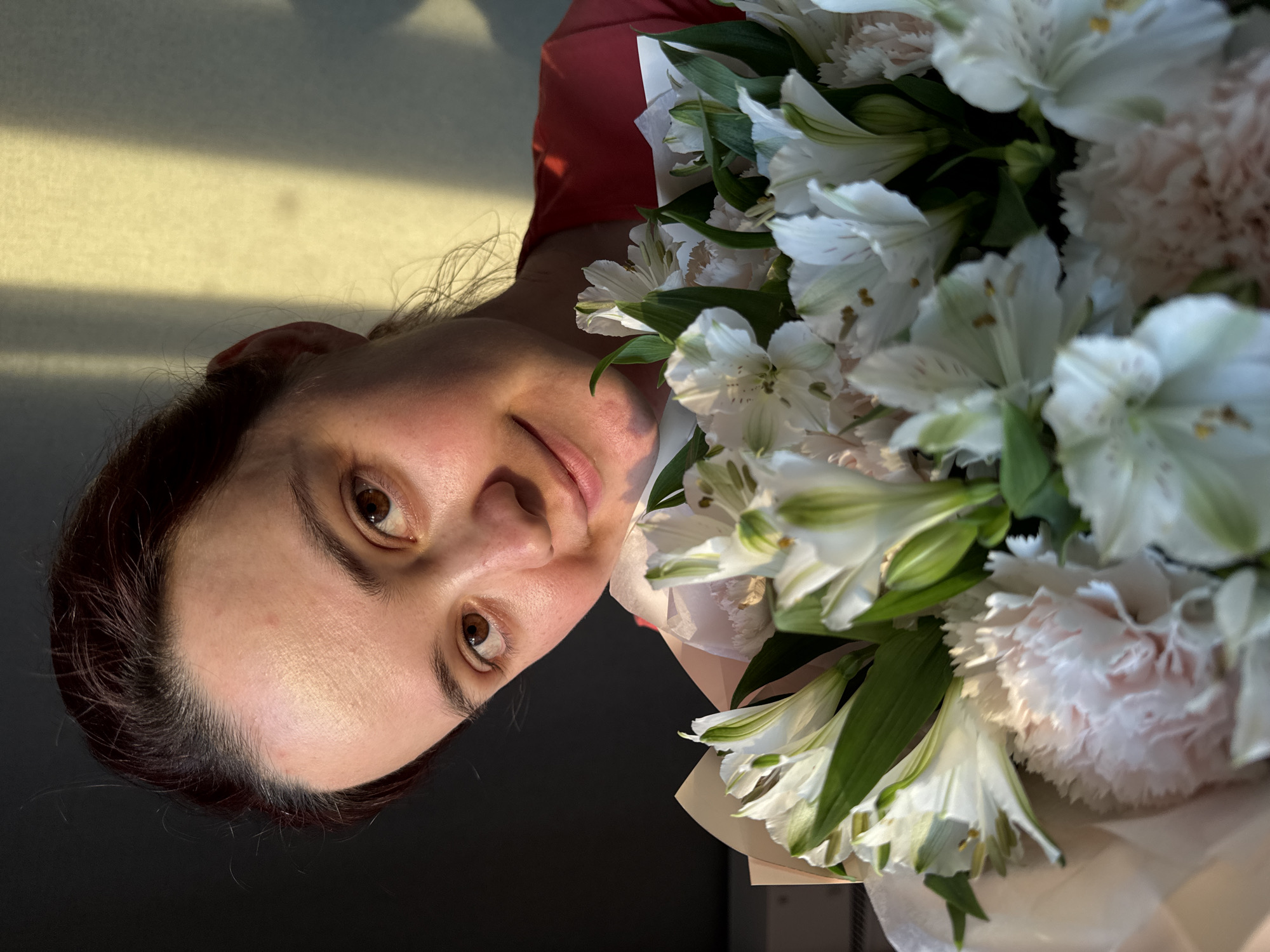 The media file depicts a close-up, sideways portrait of a woman holding a bouquet of white alstroemeria and pink carnations. The woman has brown eyes and dark hair. She is wearing a red shirt. The lighting suggests it is daytime, with a sunbeam casting a shadow across the background. The background appears to be indoors, with a dark surface in the foreground and a lightly colored wall with a striped shadow pattern behind her. The overall mood is gentle and possibly serene, with the soft lighting and the presence of flowers. There is no visible text in the image. The location context provided is Izhevsk, Russia.