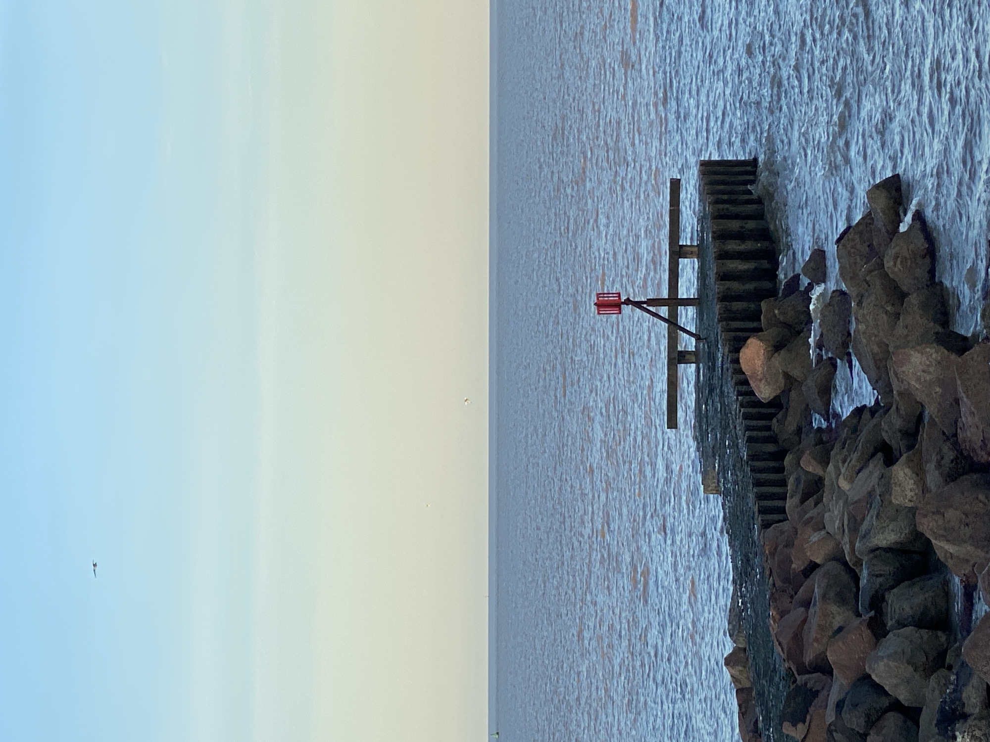 The image depicts a section of the coastline in East Suffolk, United Kingdom, likely near the sea. A series of dark, jagged rocks lines the right side of the frame, partially submerged in the water. Adjacent to the rocks, a wooden structure, possibly a pier or groyne, extends into the water, marked with a small, red navigation beacon on a post. The water stretches out towards the horizon, occupying the central portion of the image. The left side shows the sky transitioning from blue to a lighter, almost pale yellow hue. A few birds are visible in the sky. The image appears to have been rotated, as the sky is on the left rather than above.