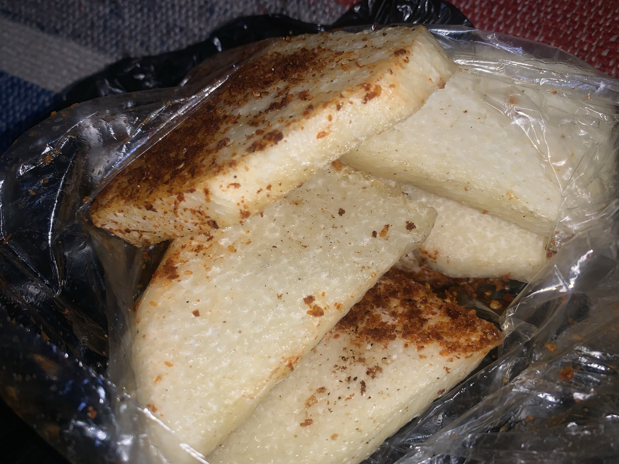 The image shows a close-up view of several pieces of a white, starchy food item, possibly plantain or yam, cut into rectangular shapes and seasoned with a reddish-brown spice blend. The food is enclosed in a clear plastic bag, and some of the pieces are layered on top of each other. The background is blurred but appears to be a patterned fabric, suggesting the food is being held or placed on a surface. The lighting is dim, creating shadows and highlighting the texture of the food and the plastic bag. There are no people visible in the image, and the scene does not provide any specific geographical markers to pinpoint its location in Rafin Par, Nigeria.