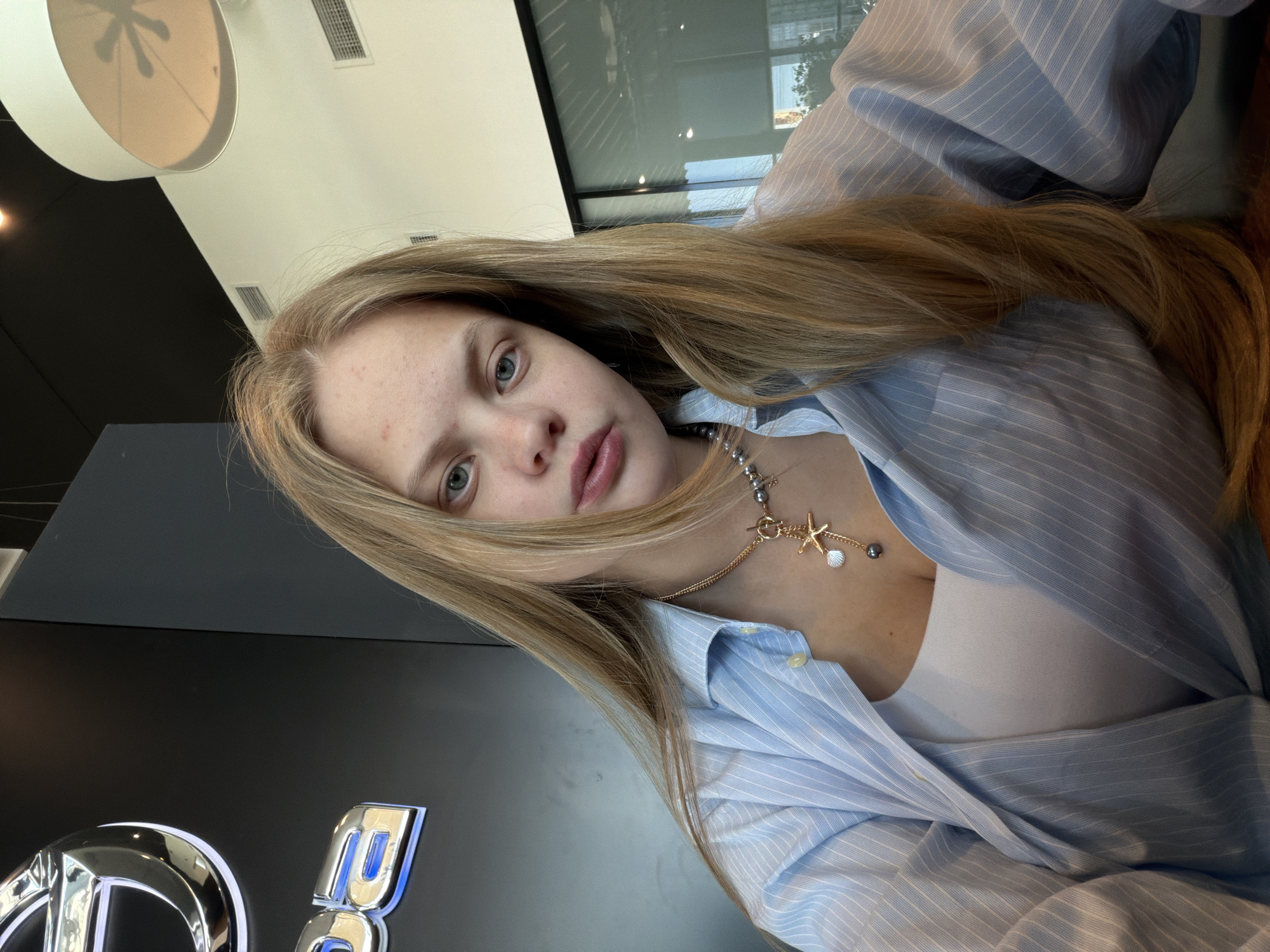 A young woman with long blonde hair and fair skin is photographed from a slightly high angle. She is wearing a light blue striped button-down shirt over a white top. The collar of the shirt is open, revealing a gold necklace with a starfish pendant and a small pearl. The background shows a modern interior with black walls and a lit logo. The location appears to be in Izhevsk, Russia.