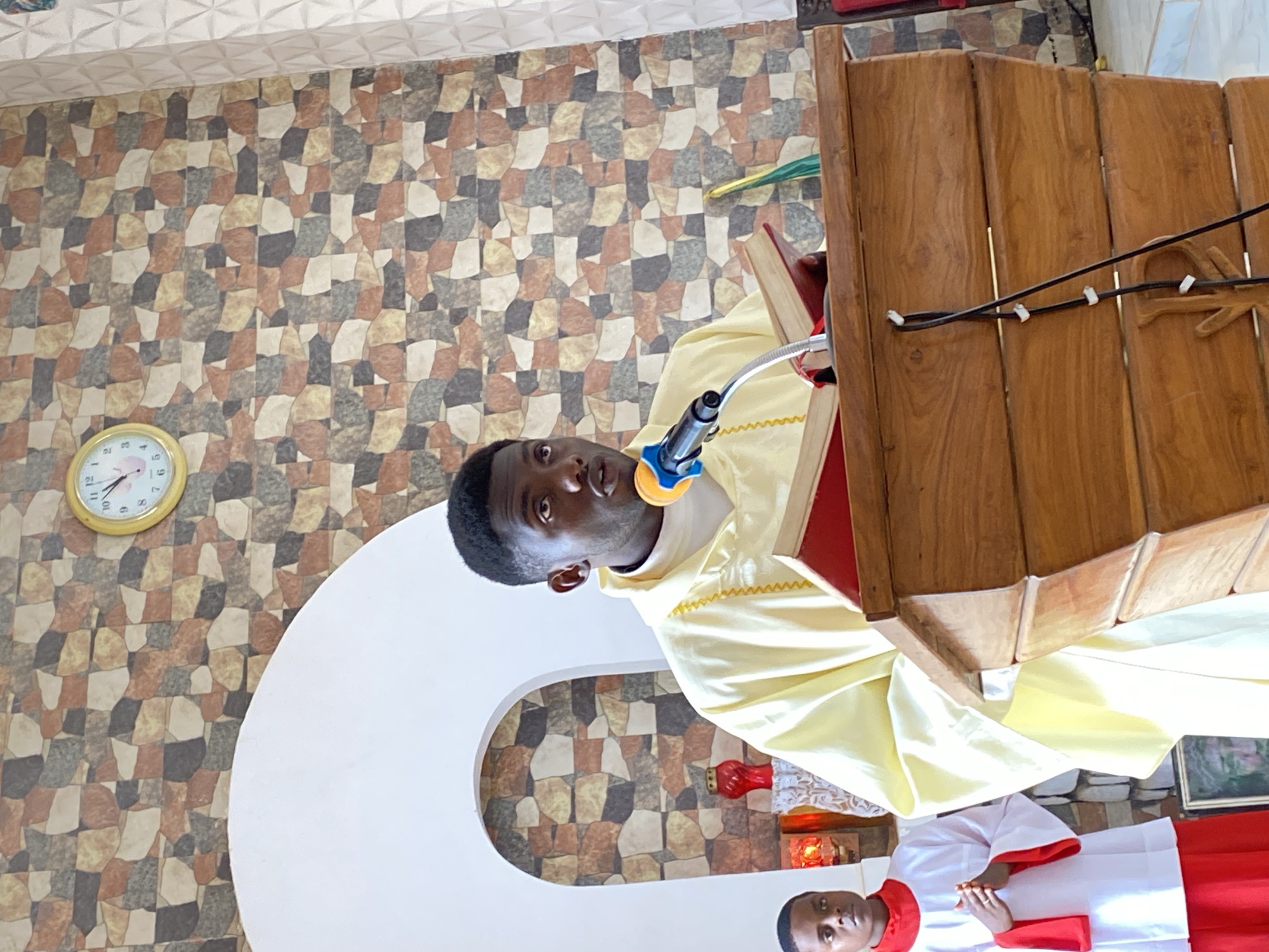 A man in a cream-colored robe stands at a wooden lectern with a microphone, seemingly preaching. Another man in a white and red robe stands below him. The background consists of a stone-patterned wall, a clock on the wall reading 11:58, and a white archway. There is a small red candle near the base of the archway. The location is likely a church in Kuru Dungus, Nigeria.