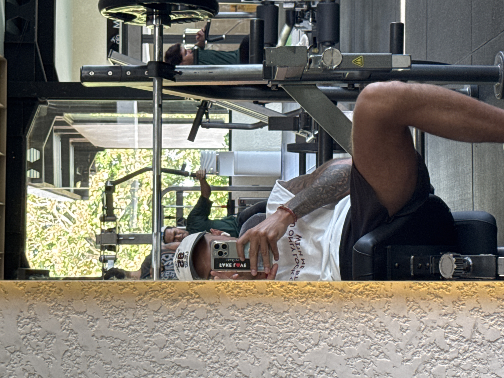 In Kuta Selatan, Indonesia, a man is taking a selfie in a gym mirror, holding a smartphone and wearing a white t-shirt and black shorts. He has tattoos on his arm and is wearing a white cap. In the reflection, another man is visible exercising with a weight machine. The gym features weightlifting equipment and a large window overlooking greenery.